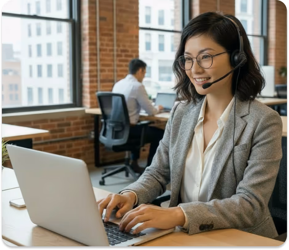 Smiling woman in glasses with headset typing on laptop in a bright office with large windows.