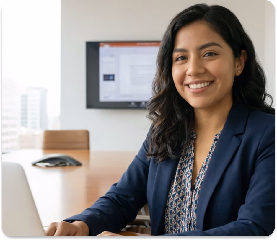 Smiling woman in a blazer sitting at a conference table with a laptop and screen in background.