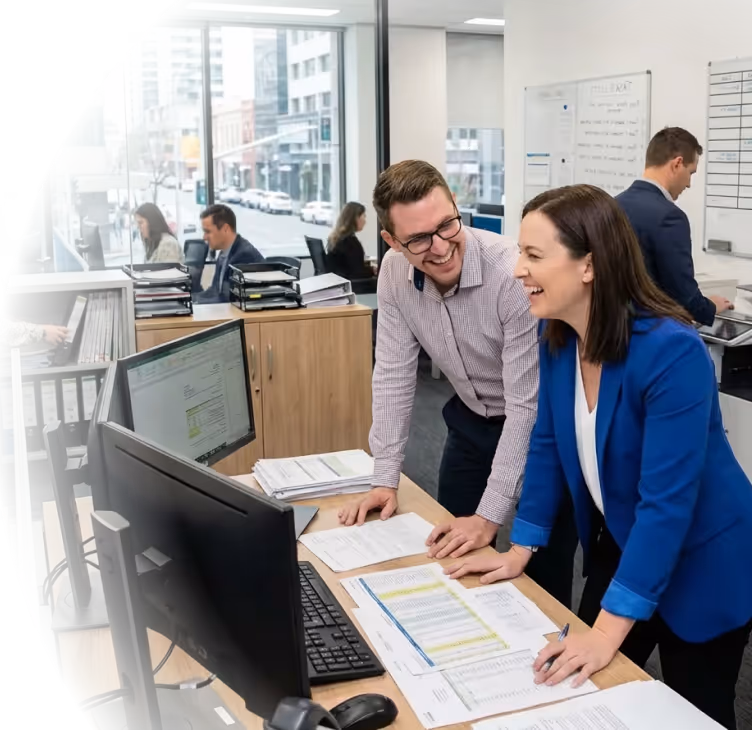 Two coworkers smiling and reviewing documents together at a desk with computer monitors in an office.