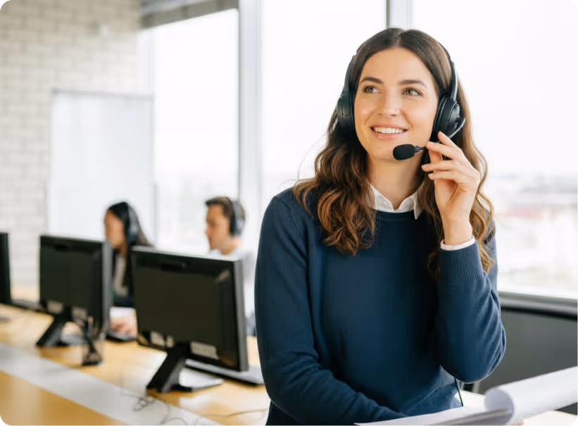 Smiling female customer service agent with headset in office, colleagues and computers in background.