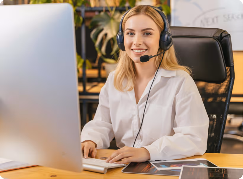 Smiling woman wearing headset working on a computer at a desk in an office with plants behind her.