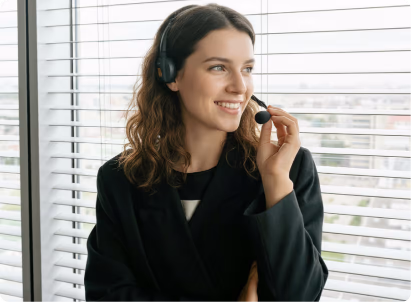 Smiling woman wearing headset and black jacket, speaking near window with city view behind blinds.
