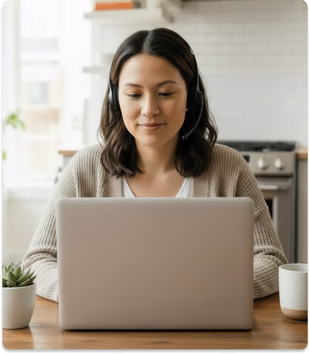 Woman wearing headset looking at laptop in kitchen with plant and coffee mug on table.