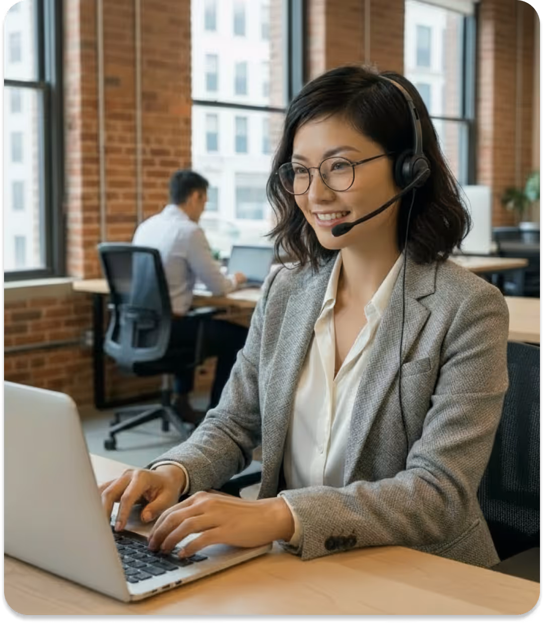 Smiling woman in glasses with headset typing on laptop in a bright office with large windows.