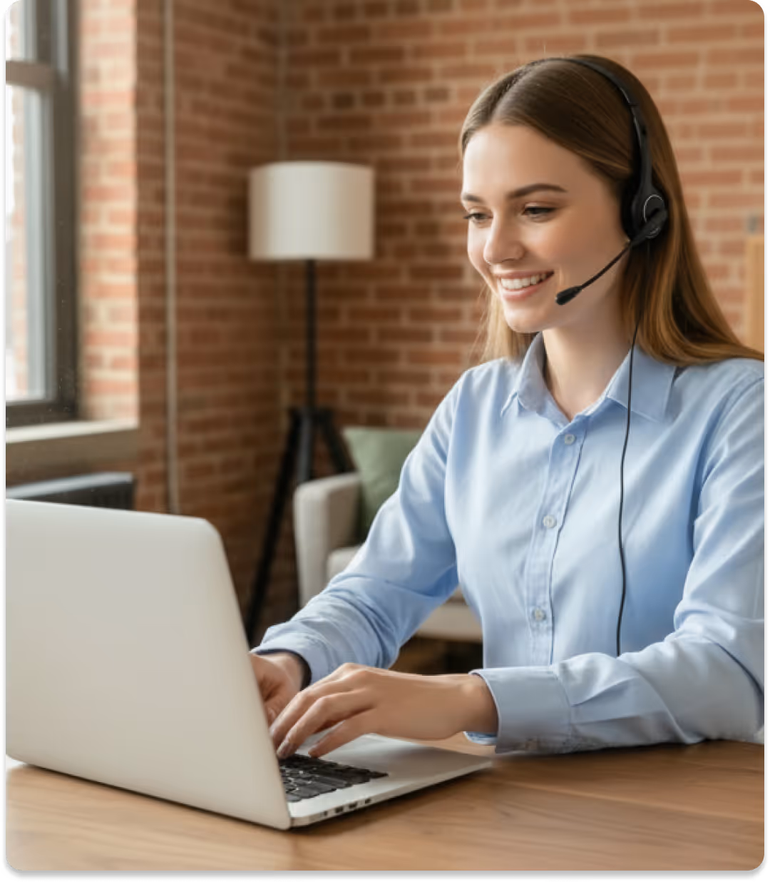 Smiling woman wearing headset typing on laptop at a wooden table in a brick-walled room.