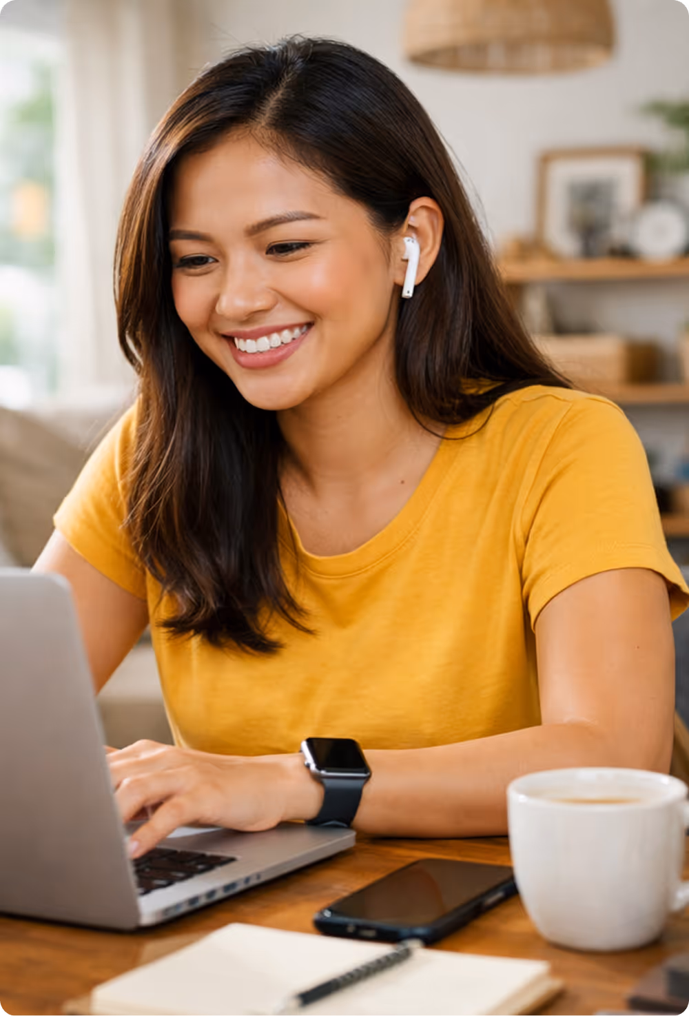 Smiling woman in a yellow shirt using a laptop with wireless earbuds, smartwatch, smartphone, notebook, and coffee mug on the table.