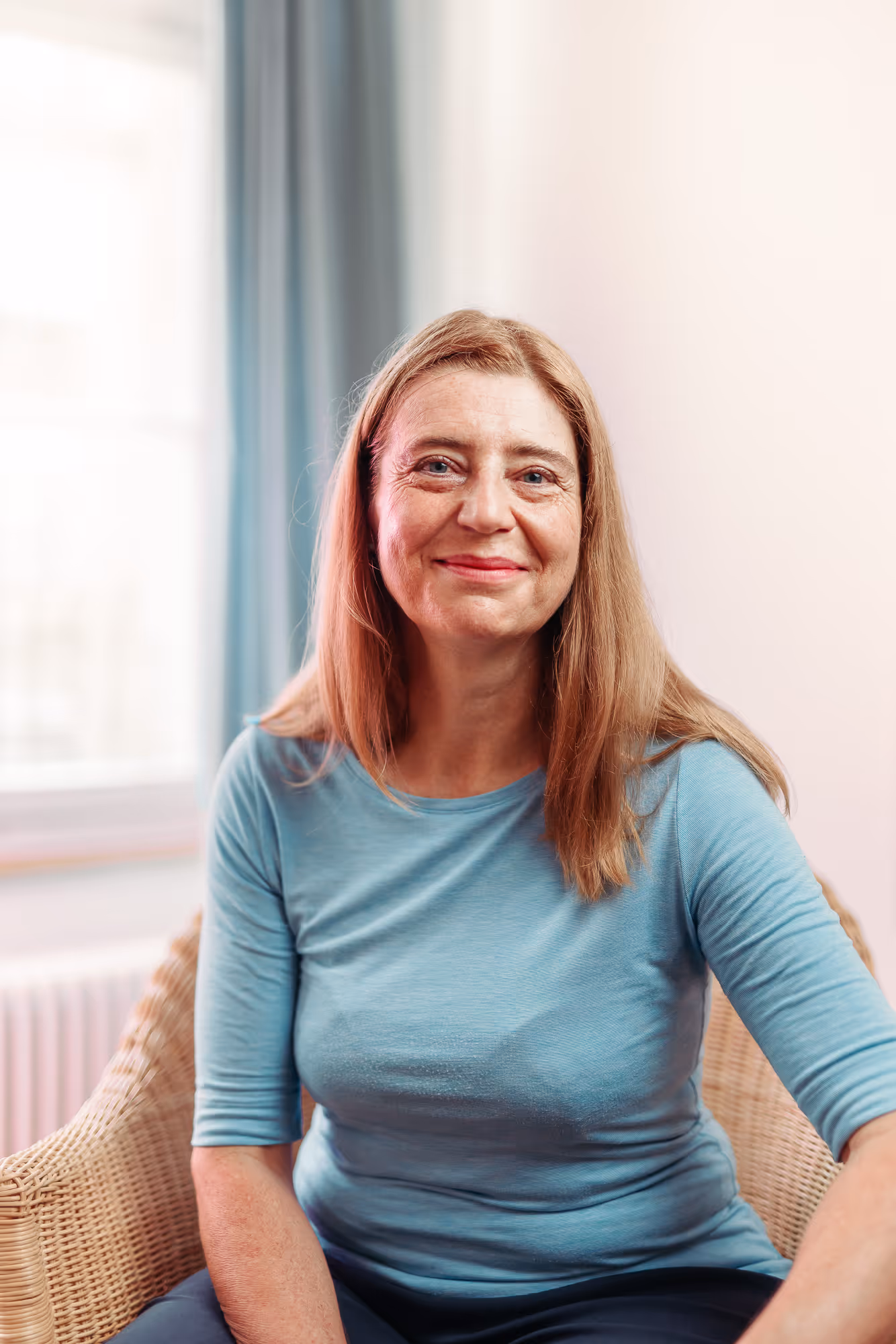 Lächelnde Frau mittleren Alters mit langen blonden Haaren, die in einem Korbsessel sitzt und ein hellblaues Oberteil trägt.