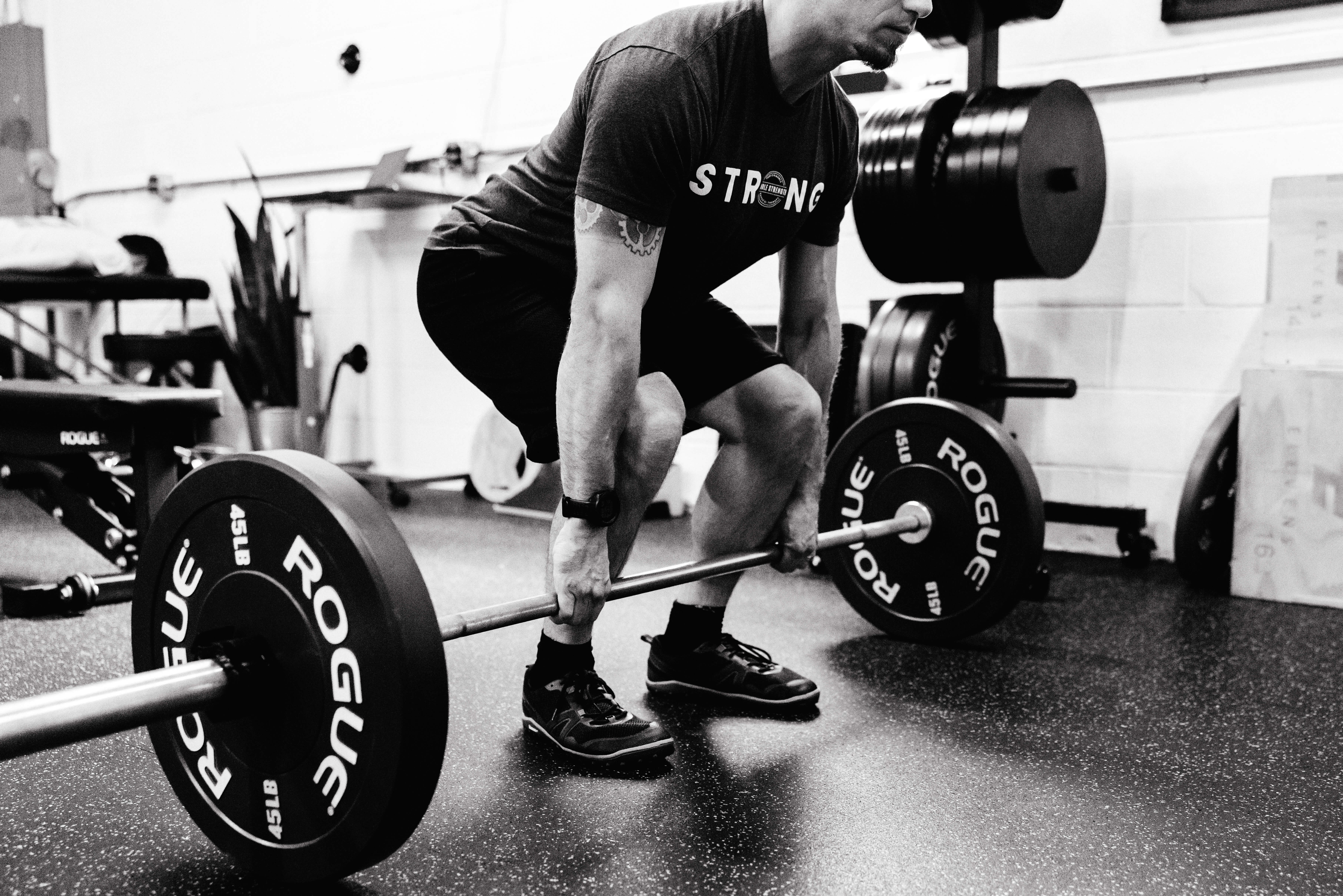 Man wearing a 'STRONG' t-shirt lifting a barbell with 45 lb Rogue weight plates in a gym.