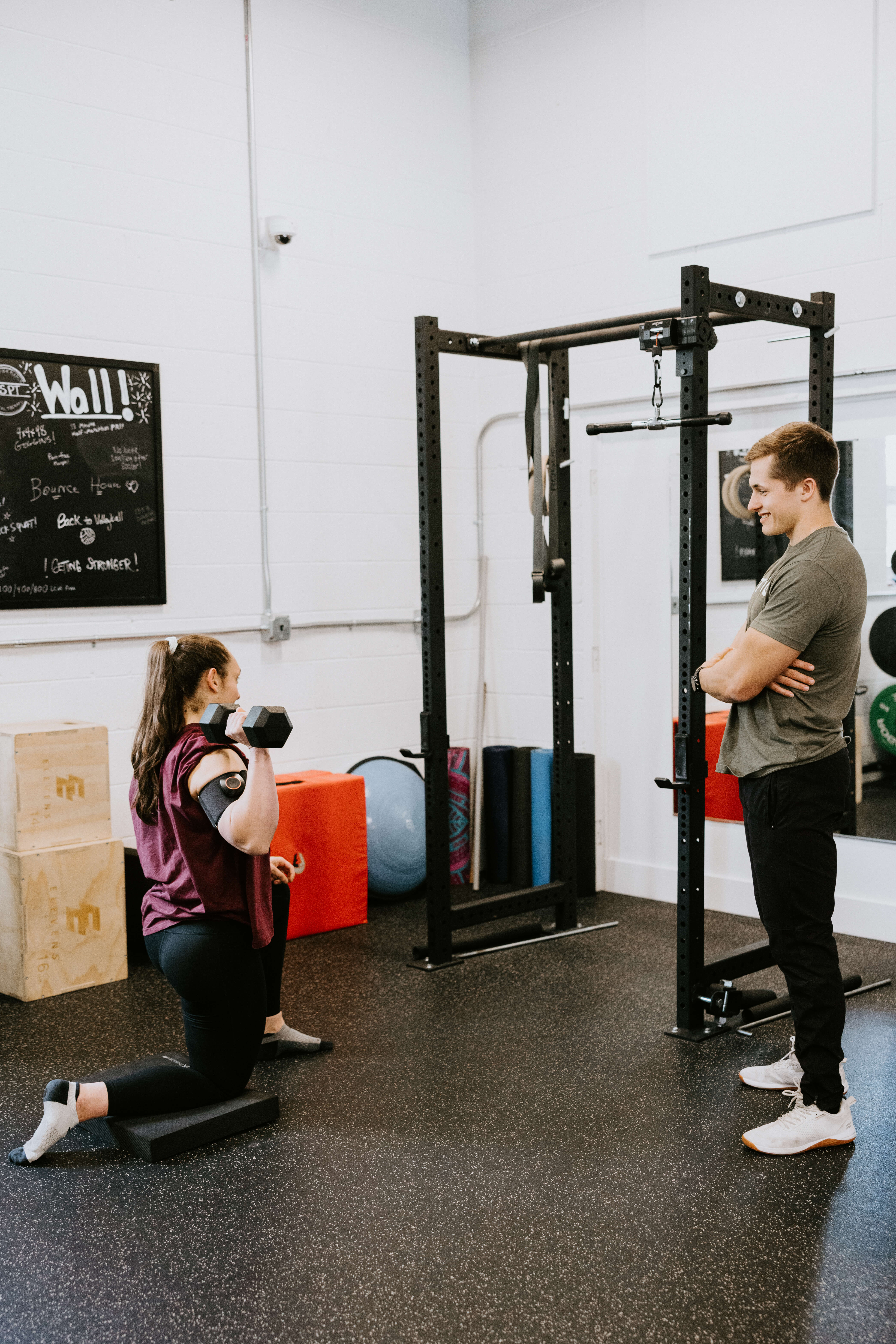 Woman kneeling on a foam pad lifting a dumbbell with a coach standing nearby in a gym.