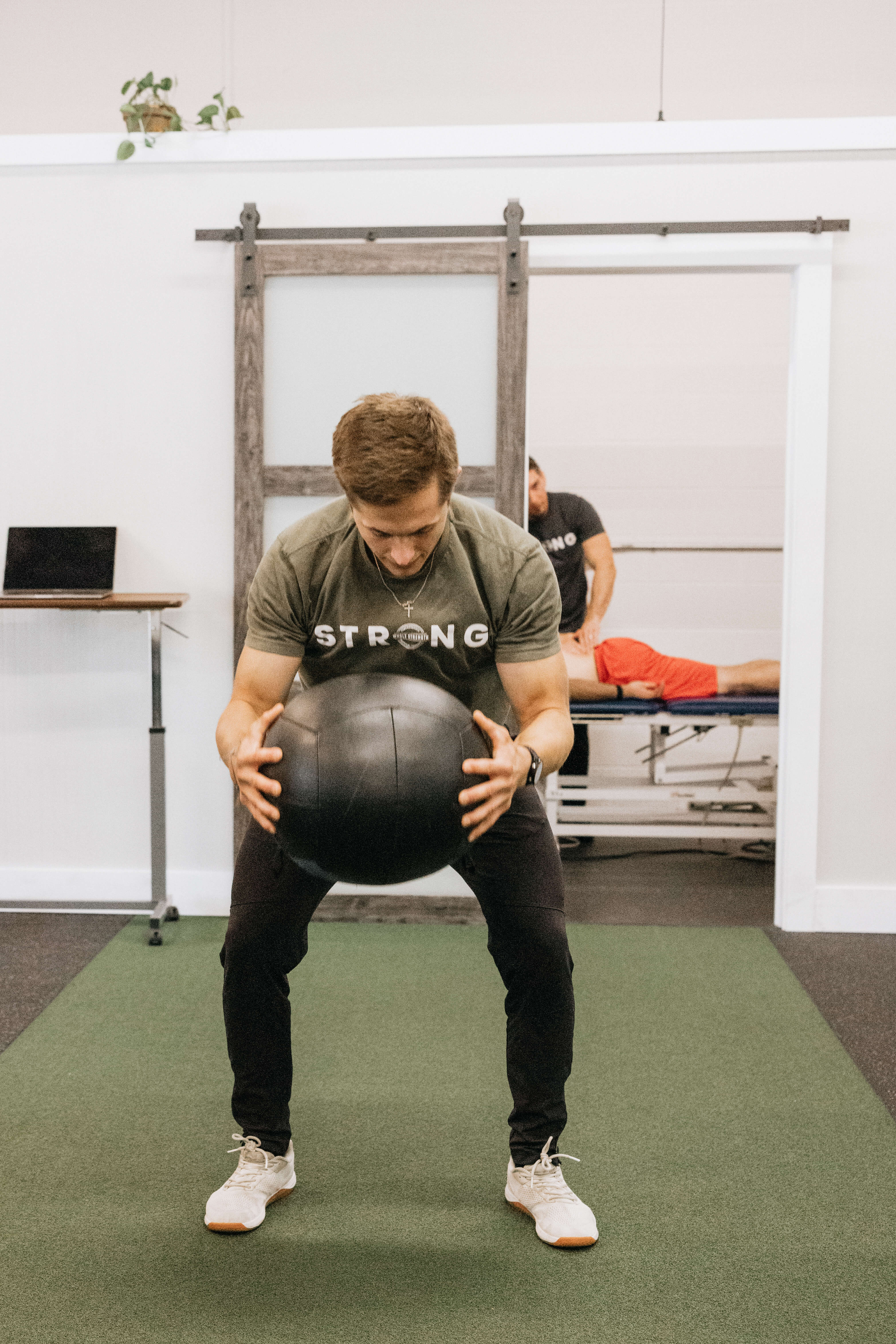 Man in green shirt and black pants holding a black exercise ball in a gym setting with another person lying on a treatment table in the background.