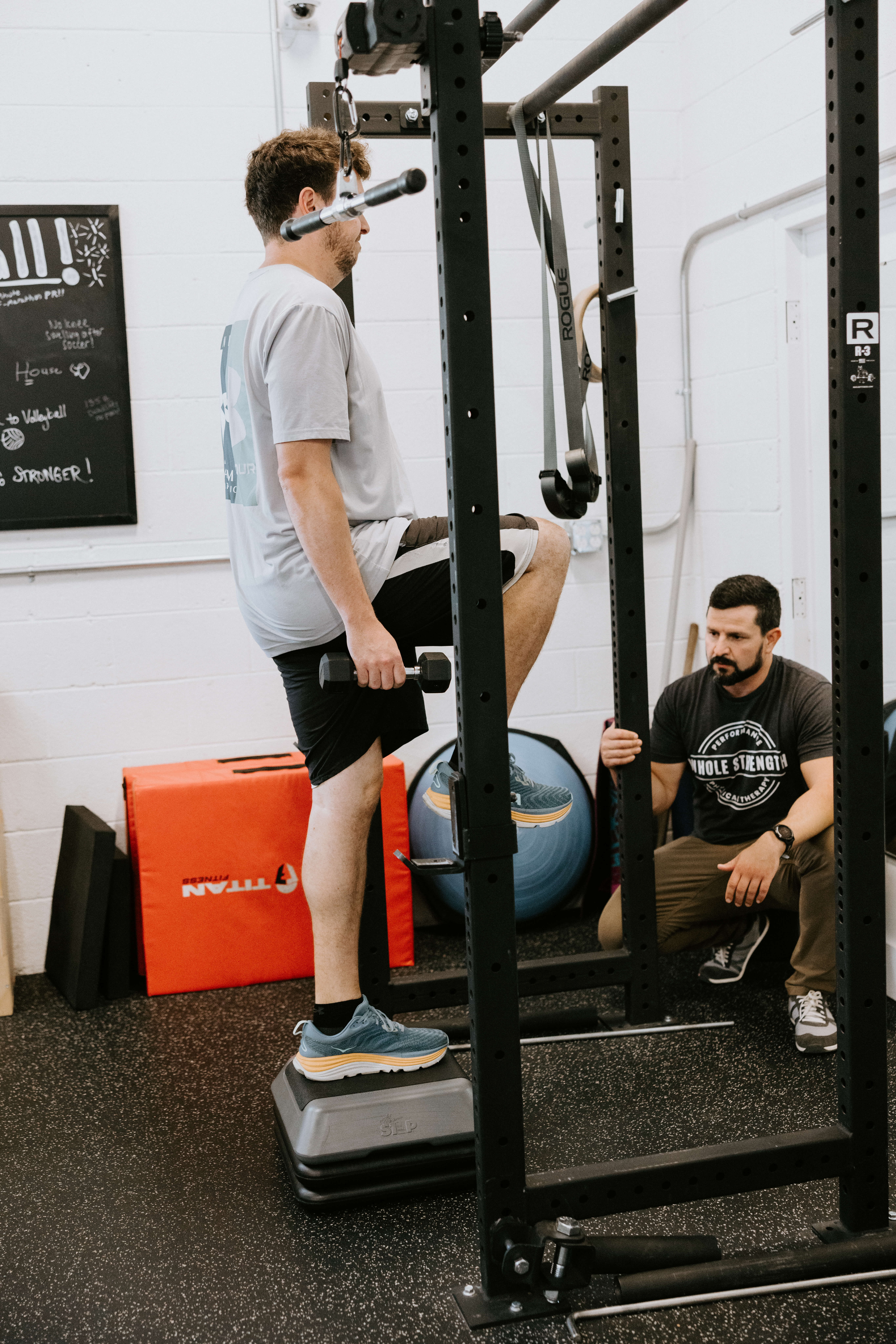 Man performing step-up exercise holding dumbbell while trainer observes in a gym.