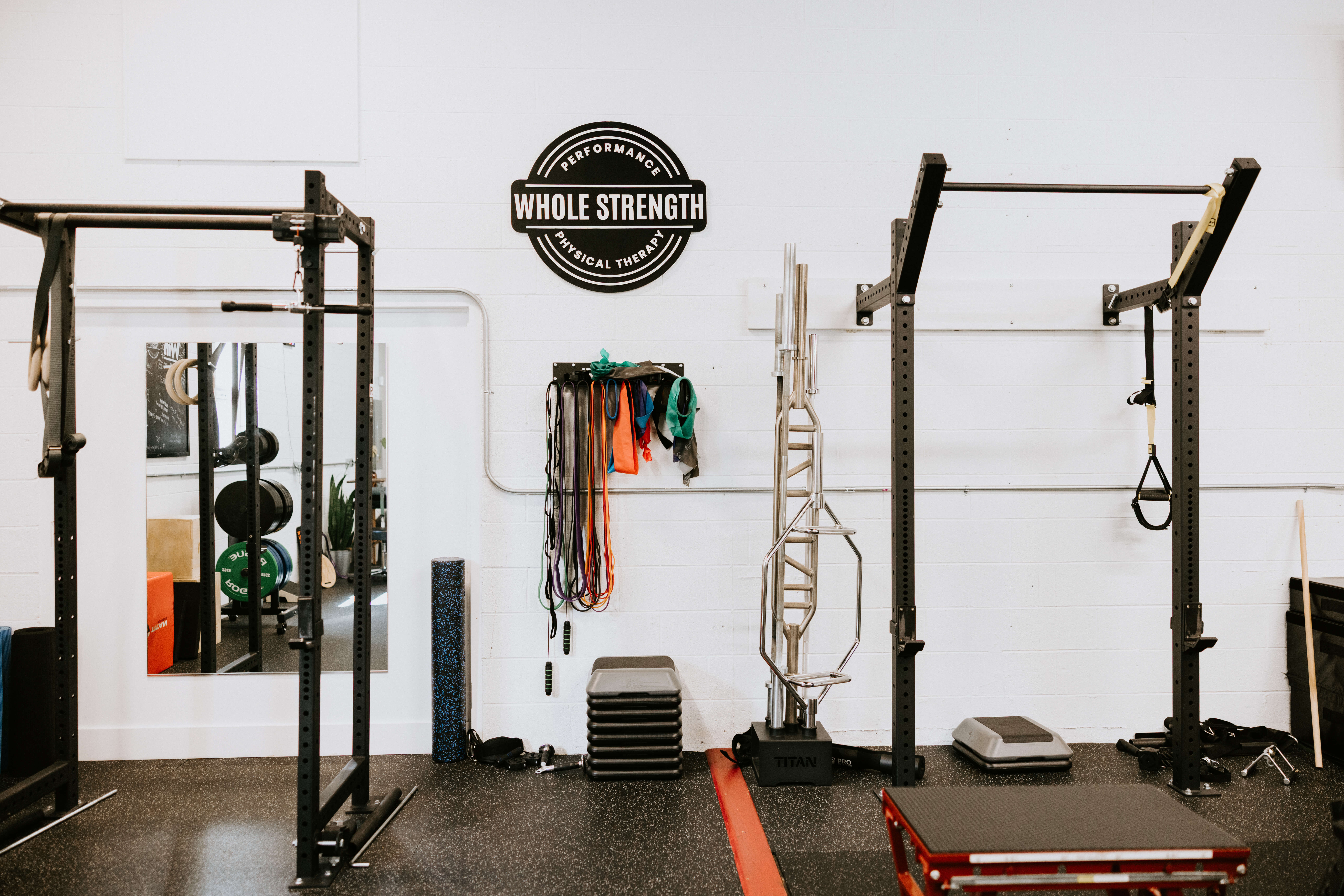 Gym area with exercise racks, resistance bands, weights, step platforms, and a sign reading Whole Strength Performance Physical Therapy on a white wall.