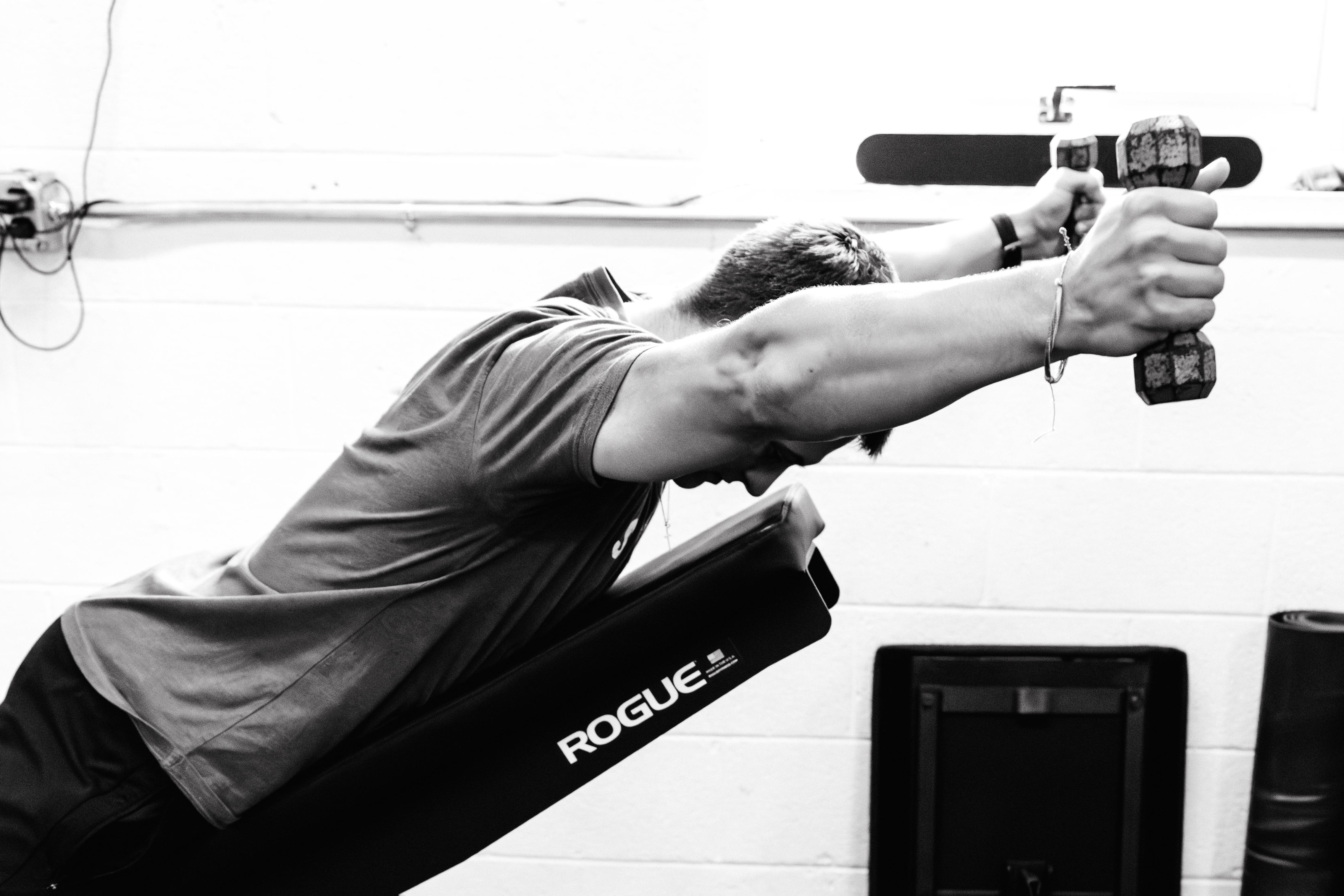 Black and white photo of man laying with chest and stomach on a slanted bench  with two dumbbells raised on both sides by his face and ears. 