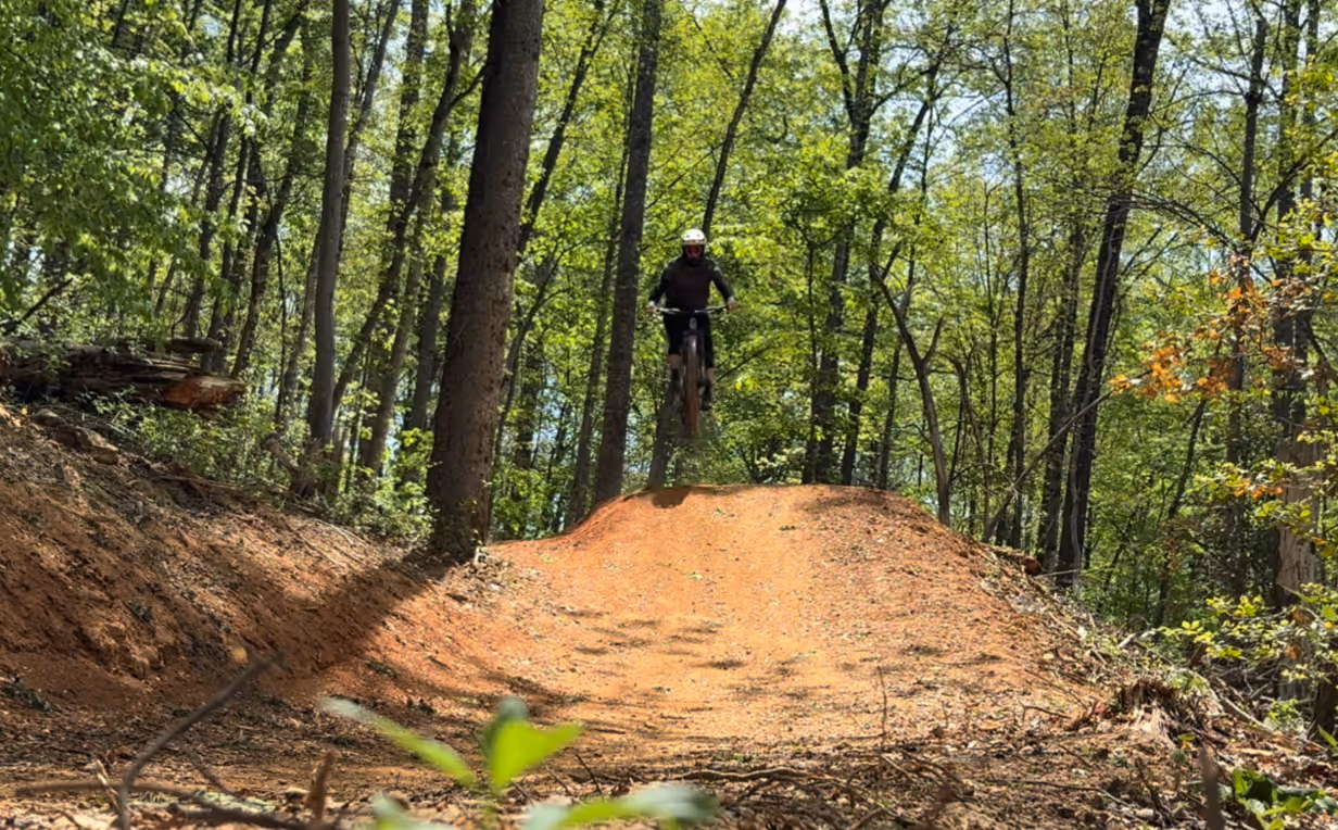 Outside view or mountain biker going over a dirt hill with trees in the background. 