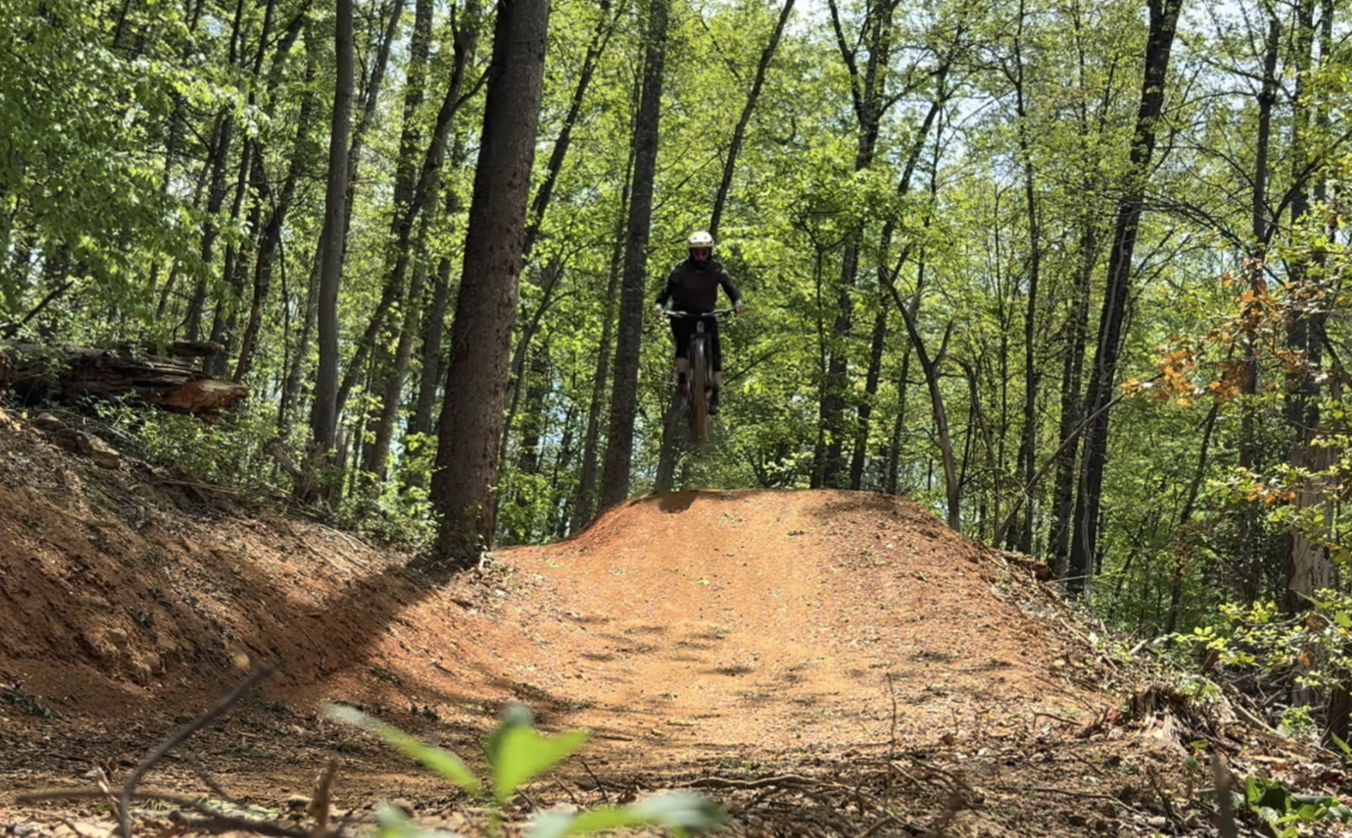 Outside view or mountain biker going over a dirt hill with trees in the background.