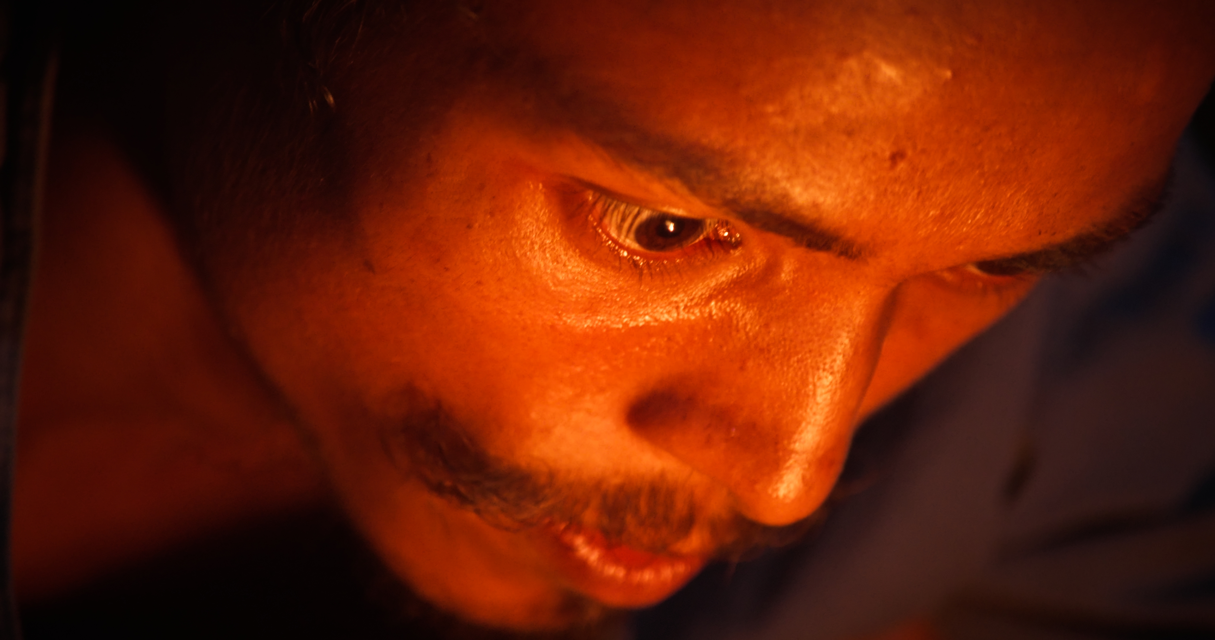 Close-up of Tikaram looking down, illuminated by warm orange light.