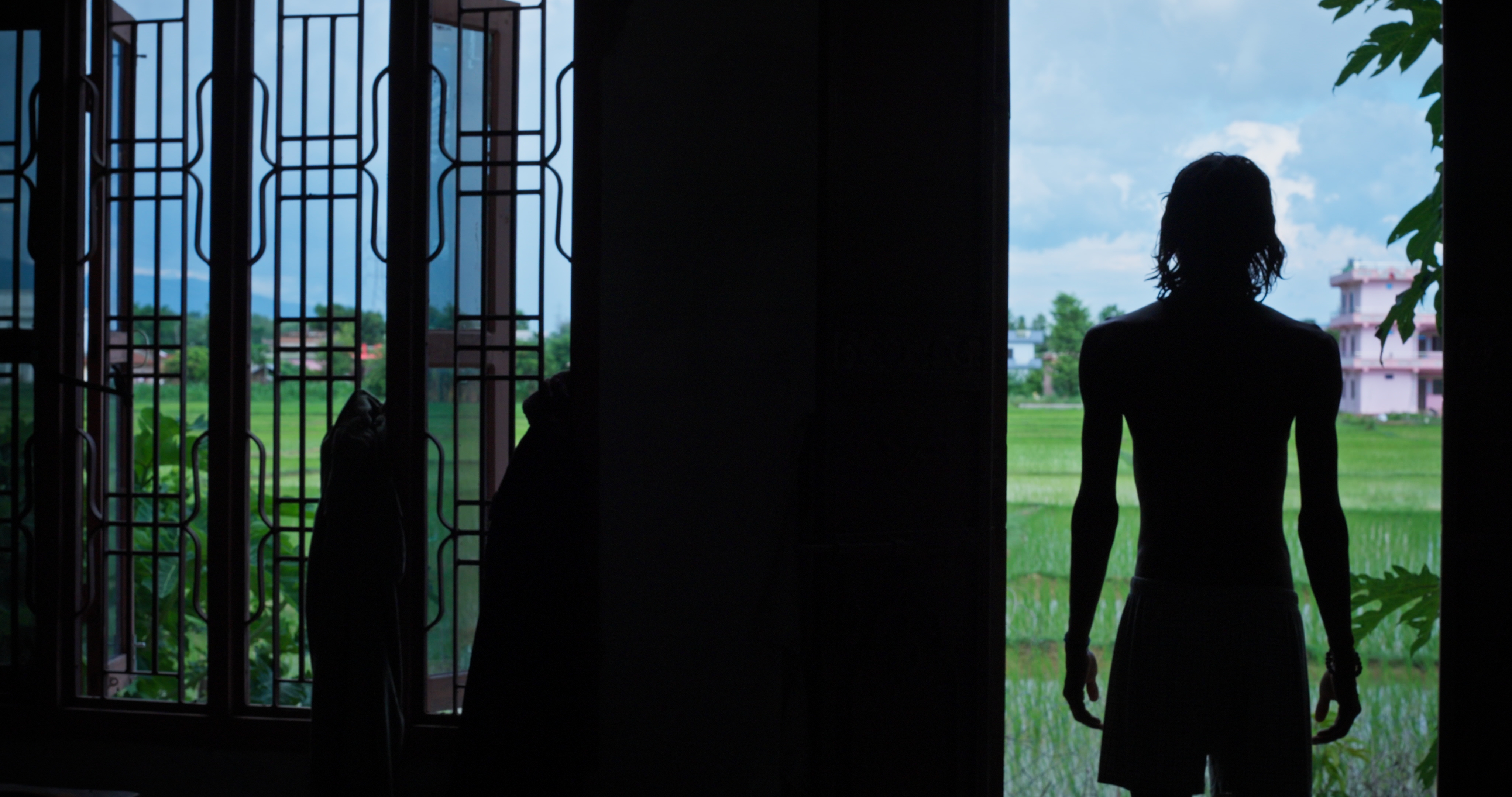 Silhouette of Tikaram standing in a doorway looking out at green fields and a pink building under a blue sky.