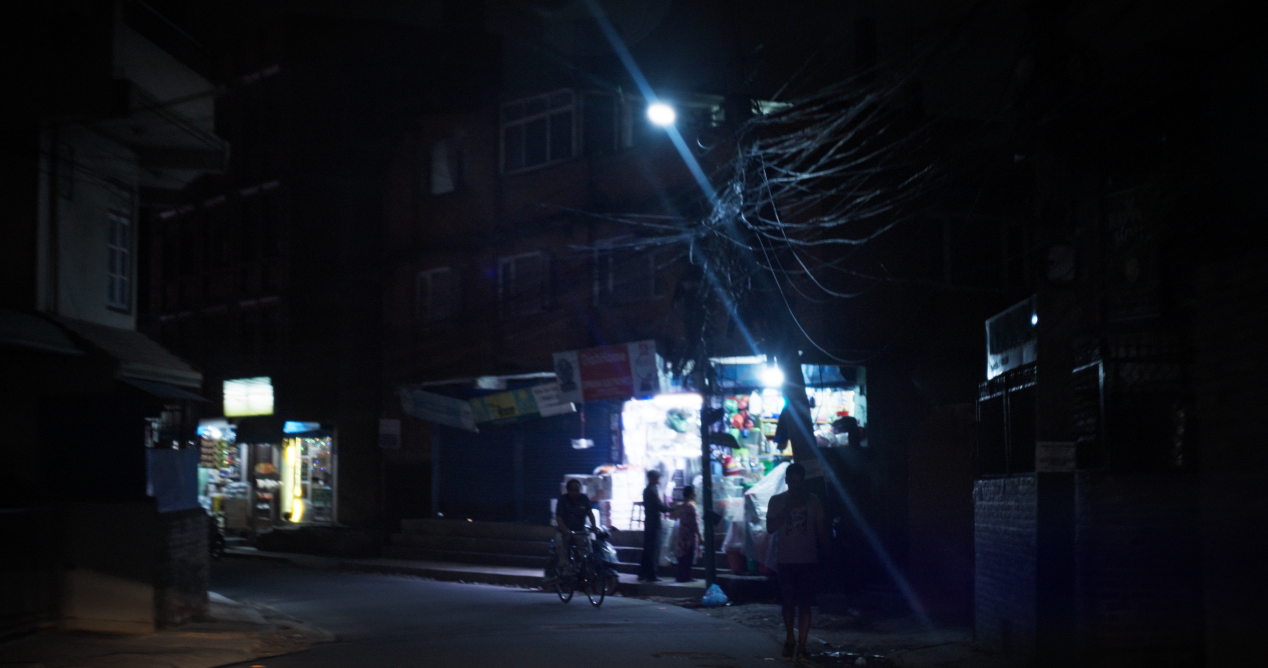 Dimly lit street at night with a small illuminated shop and people standing and cycling nearby under a streetlight.