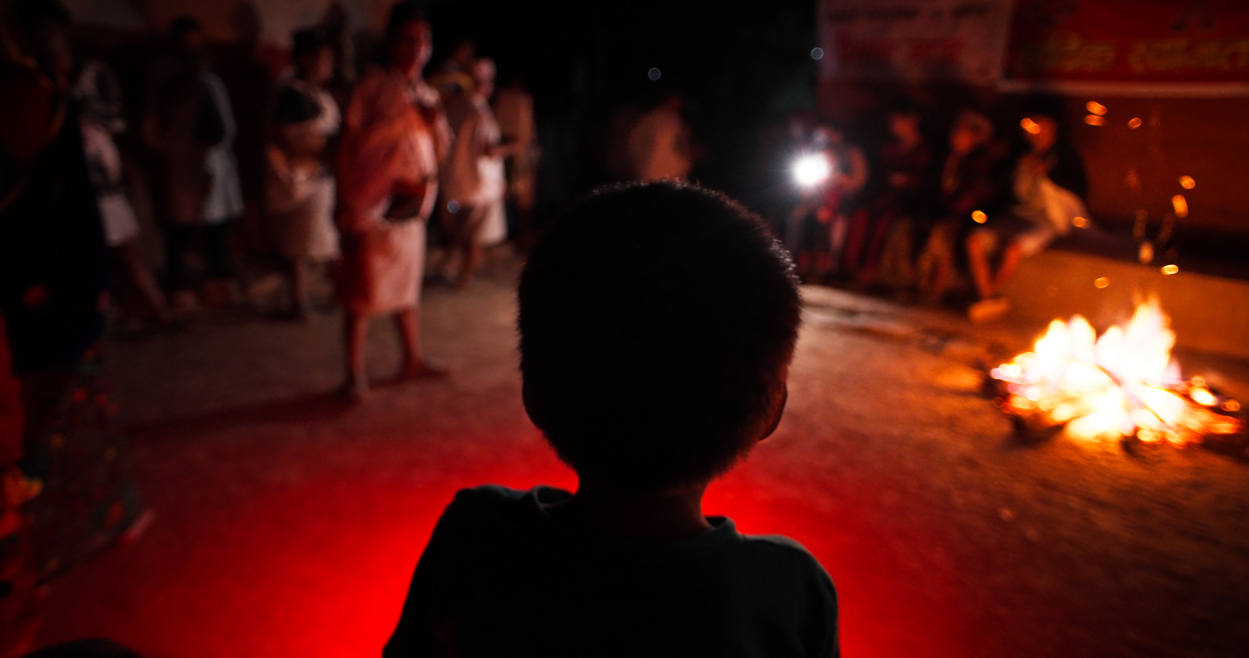 Silhouette of a child facing a group of people gathered around a fire at night.