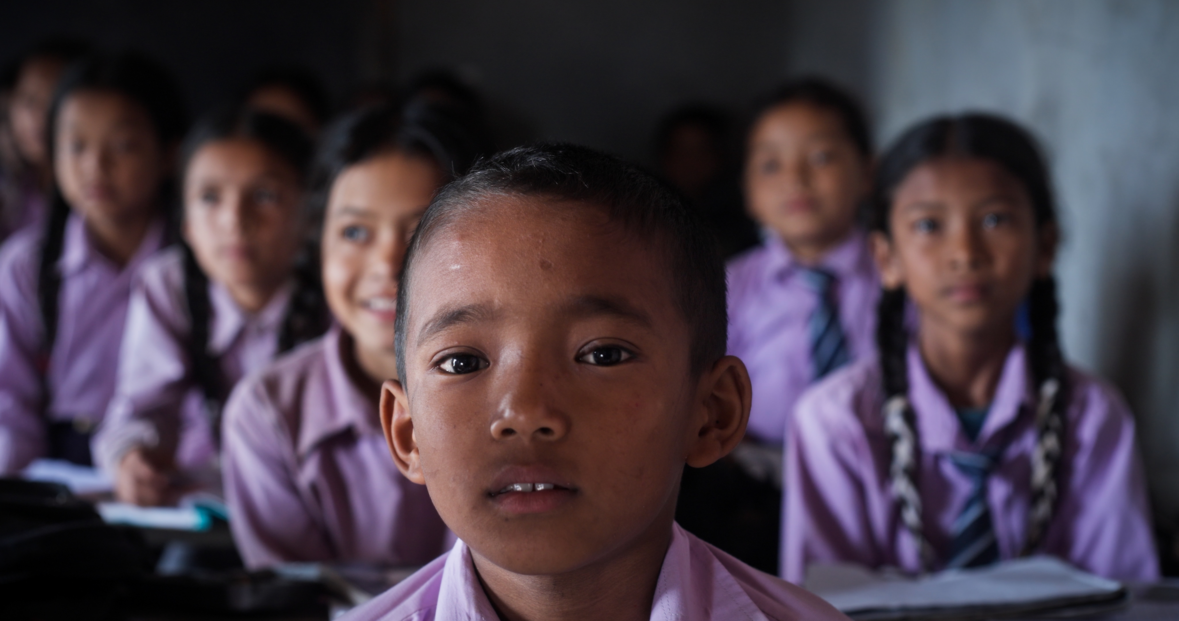 Boy in purple school uniform with classmates in the background inside a classroom.
