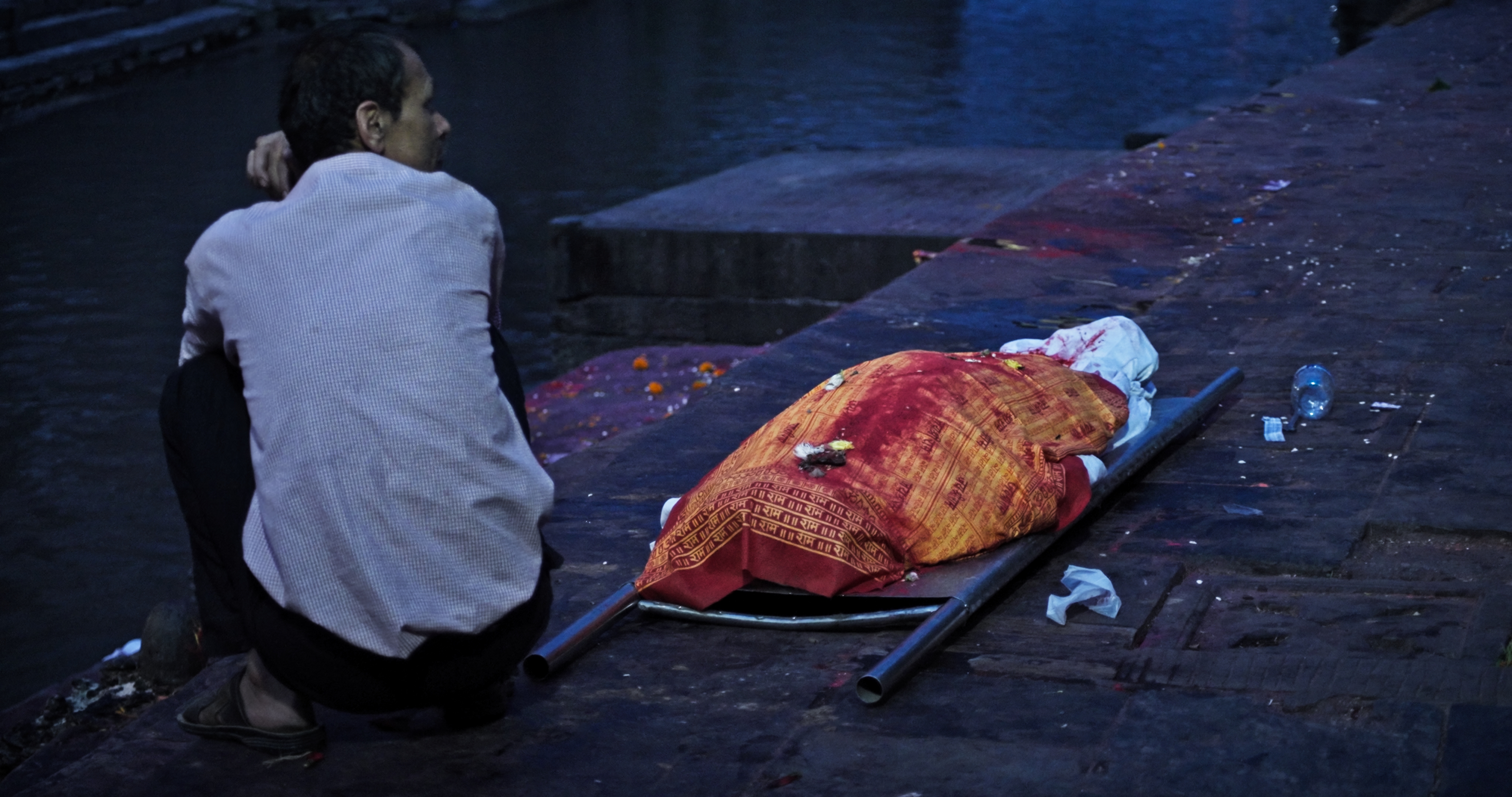 Man sitting beside a body wrapped in a red and orange cloth laid on a stretcher near a river at dusk.