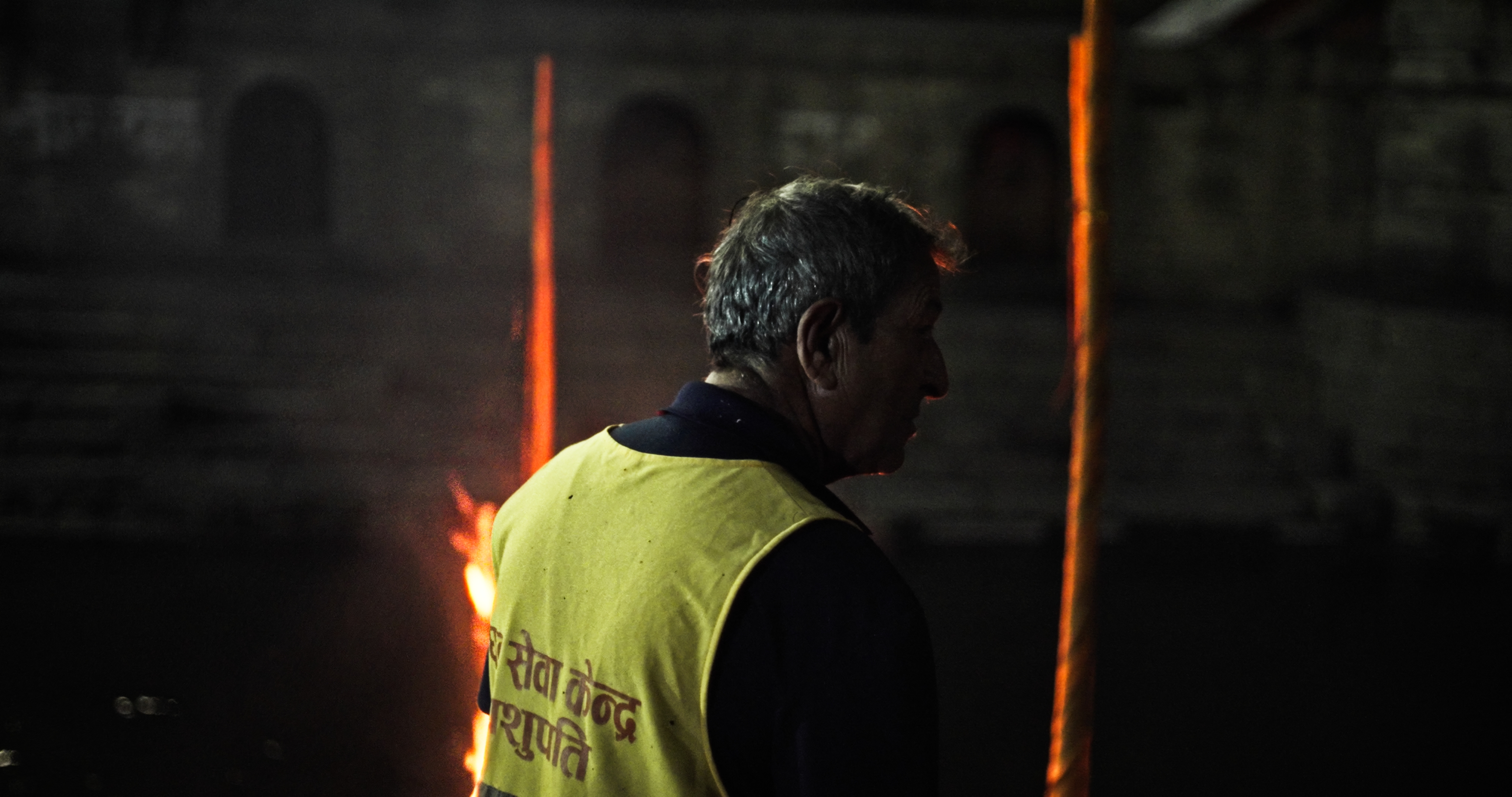Man in a yellow vest standing in front of a burning body. 