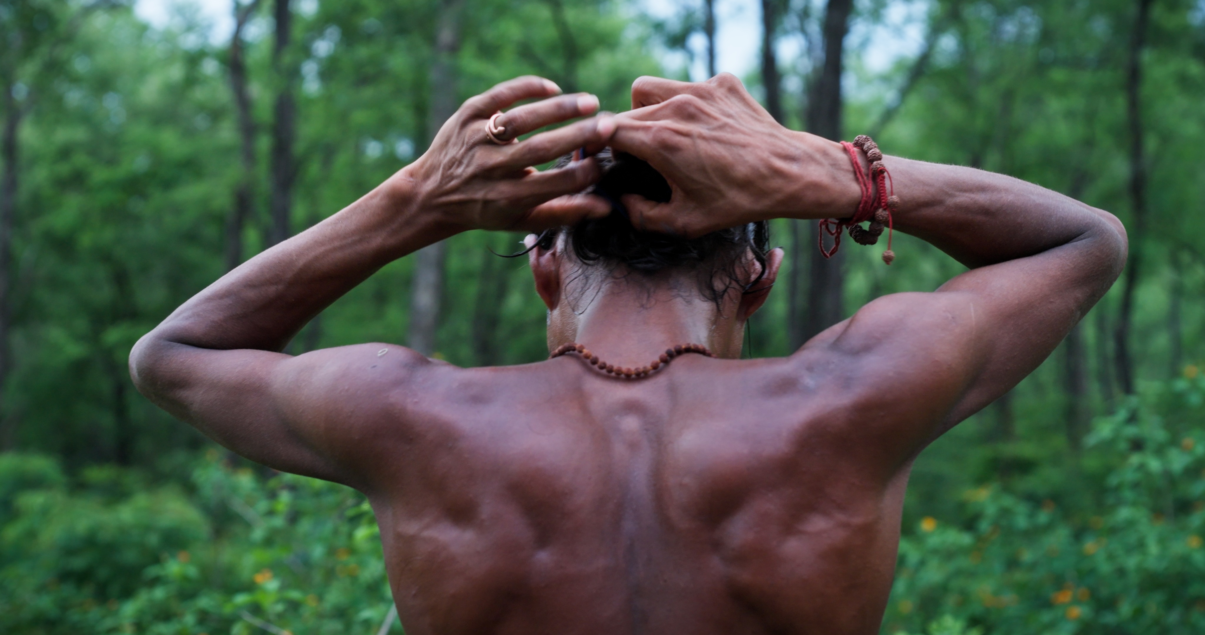 Tikaram adjusting his hair with both hands in a dense green forest.