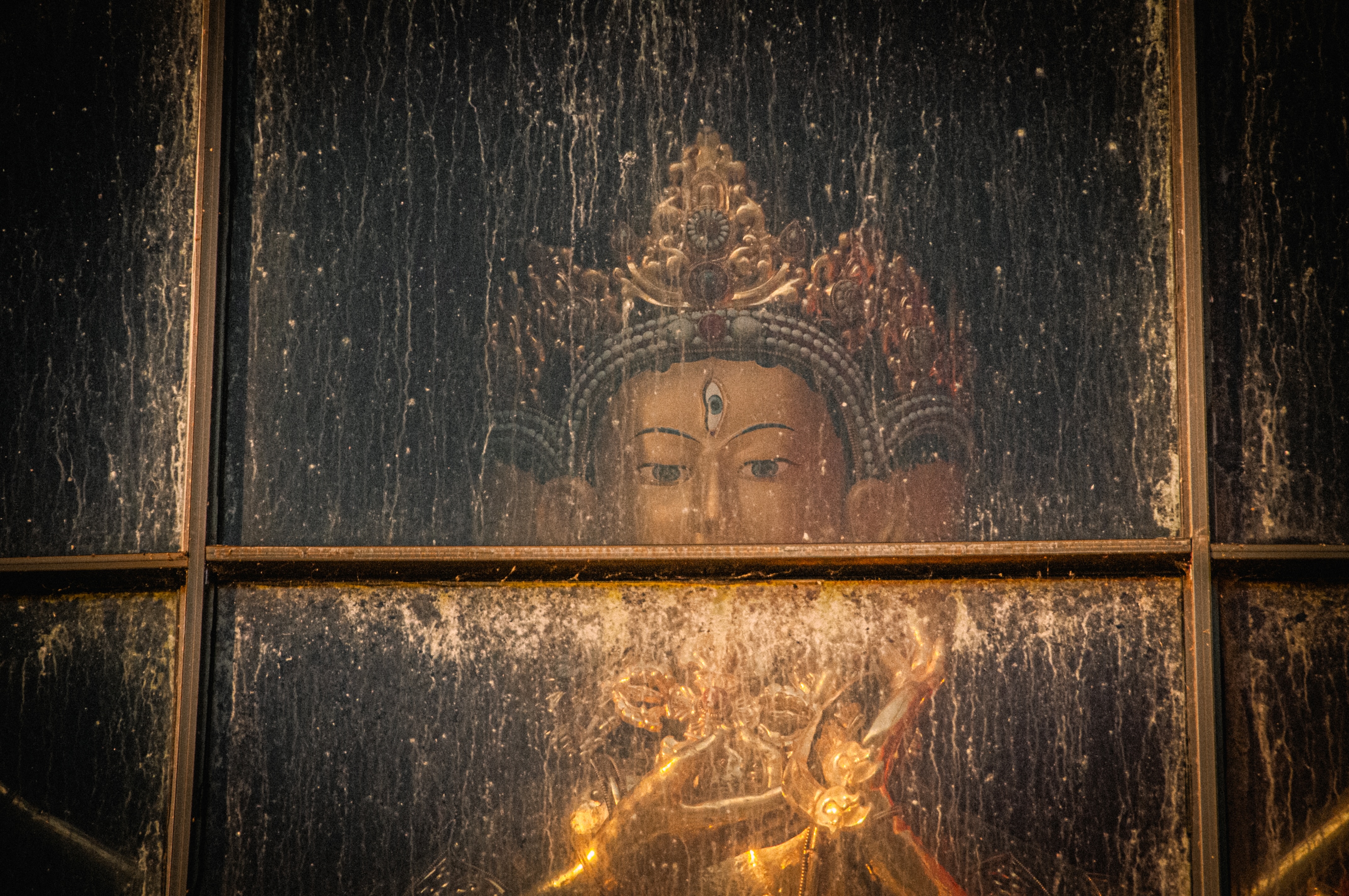 Ornate statue of a deity with a third eye behind a smudged and weathered glass pane.