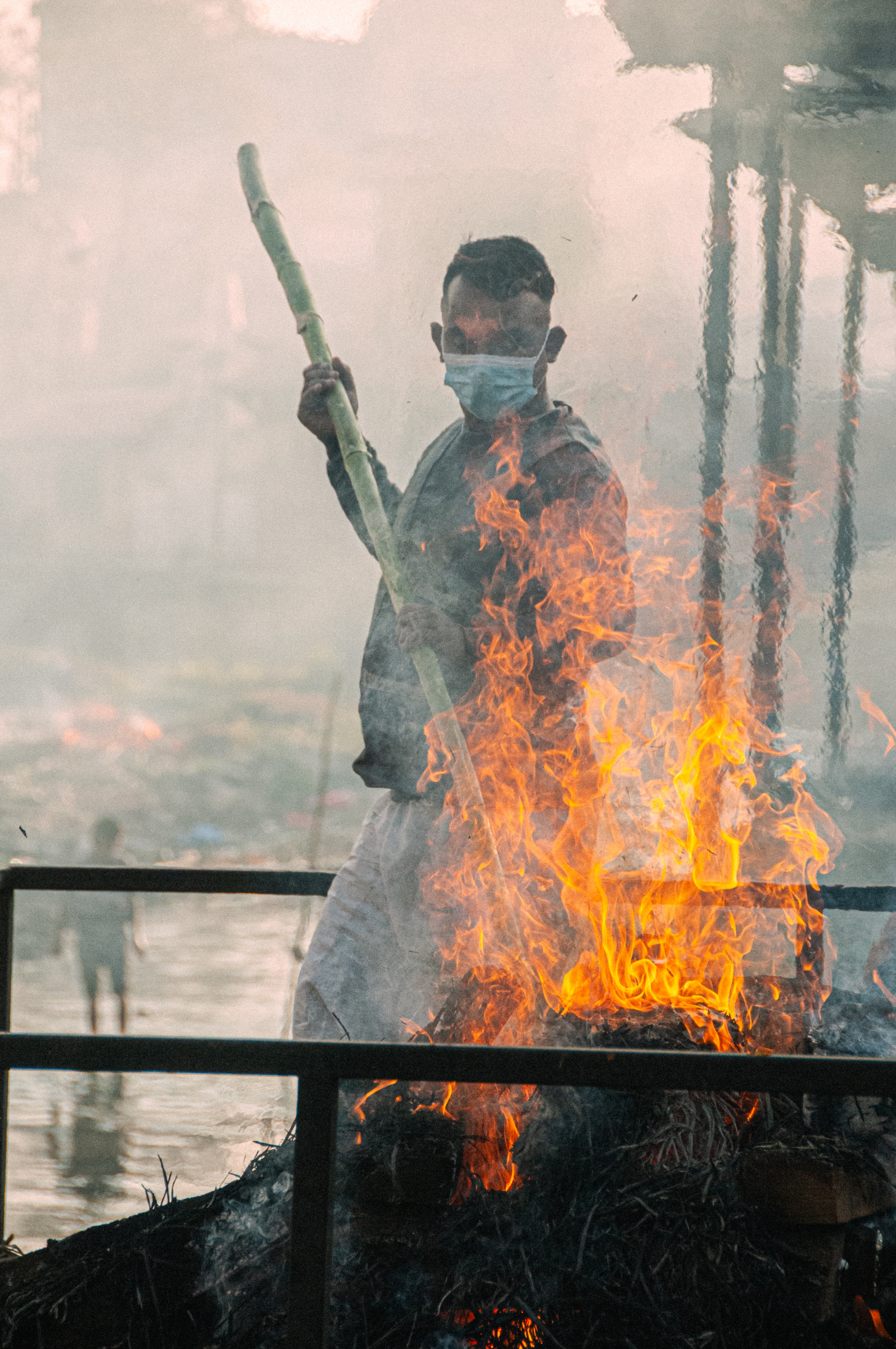 Person wearing a face mask holding a long stick near a large fire with flames and smoke rising.