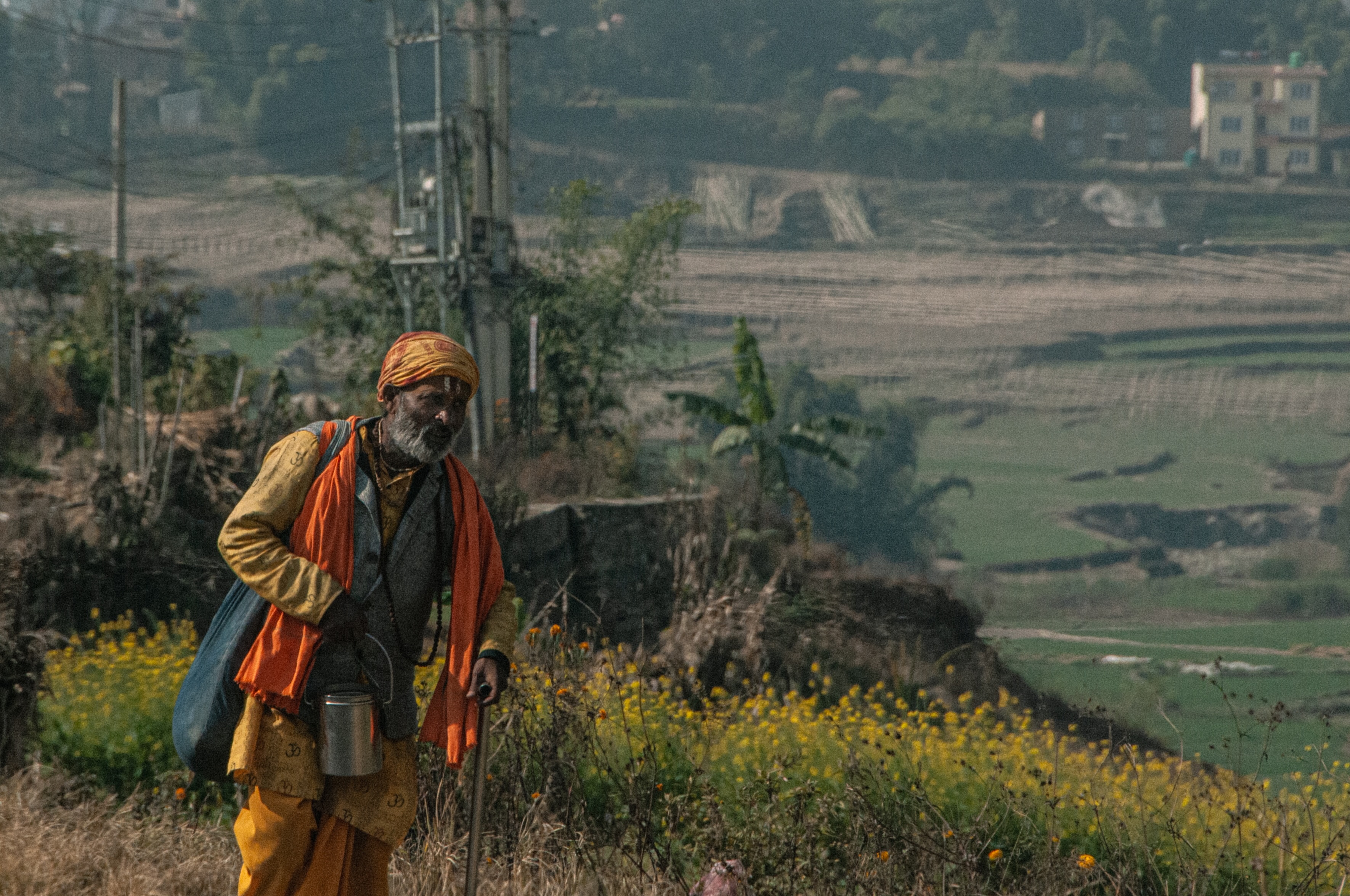 Man dressed in orange and yellow holding a metal container and walking stick in a rural landscape with yellow flowers and green fields.