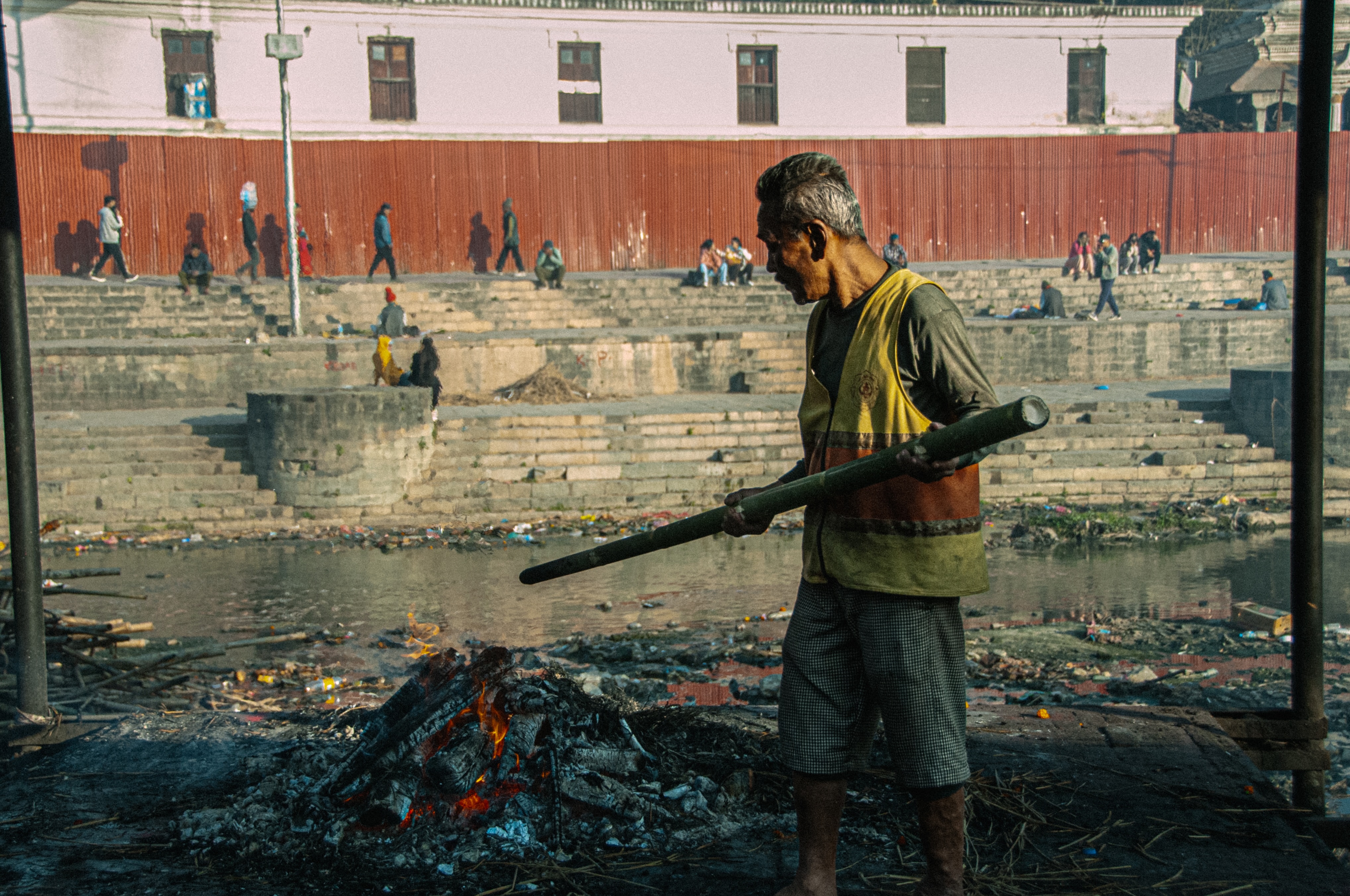 Man standing beside a burning pyre near a riverbank with stone steps and people in the background.