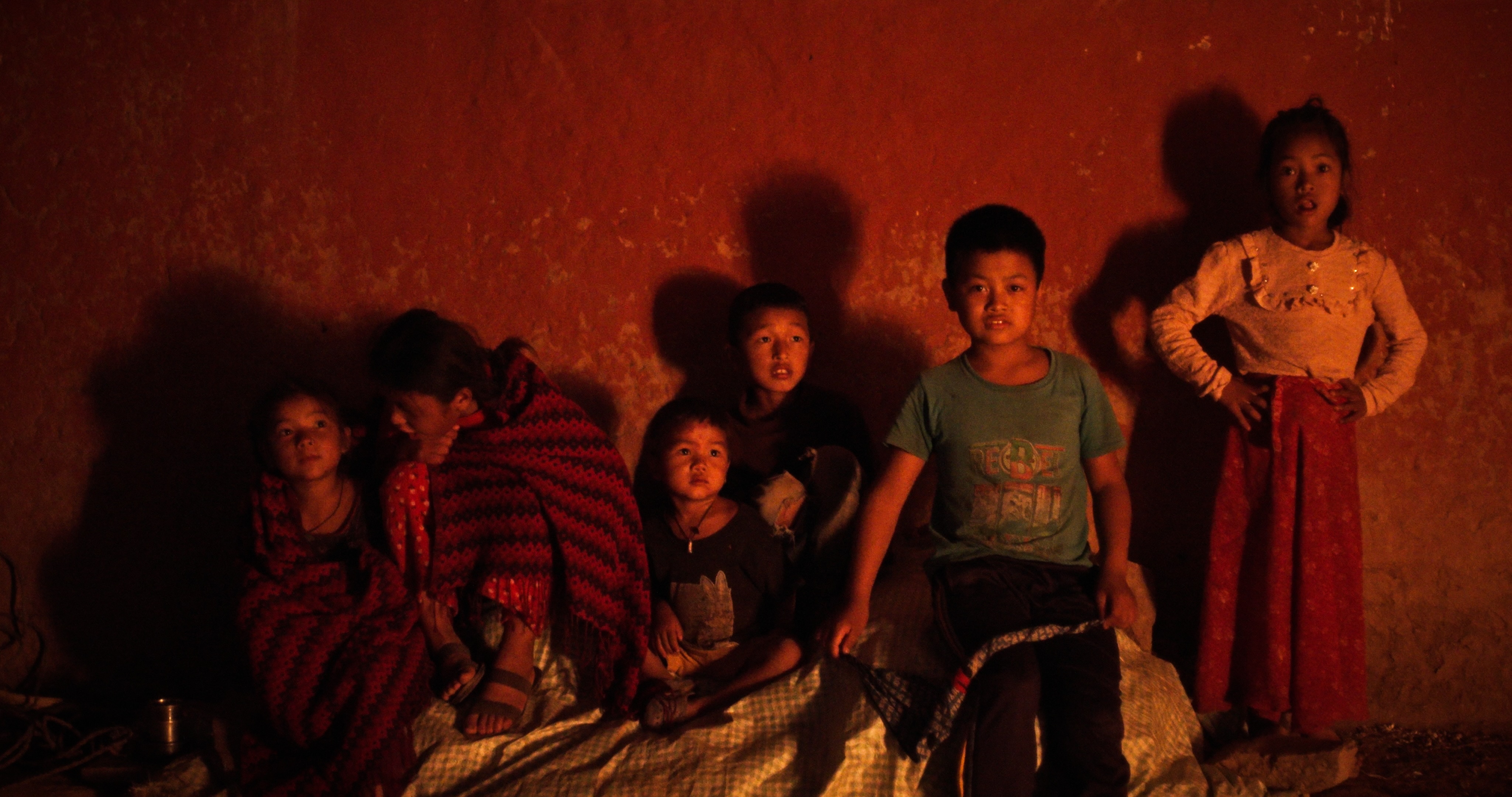 Group of six children sitting and standing against a textured reddish-brown wall, illuminated by warm, low light.