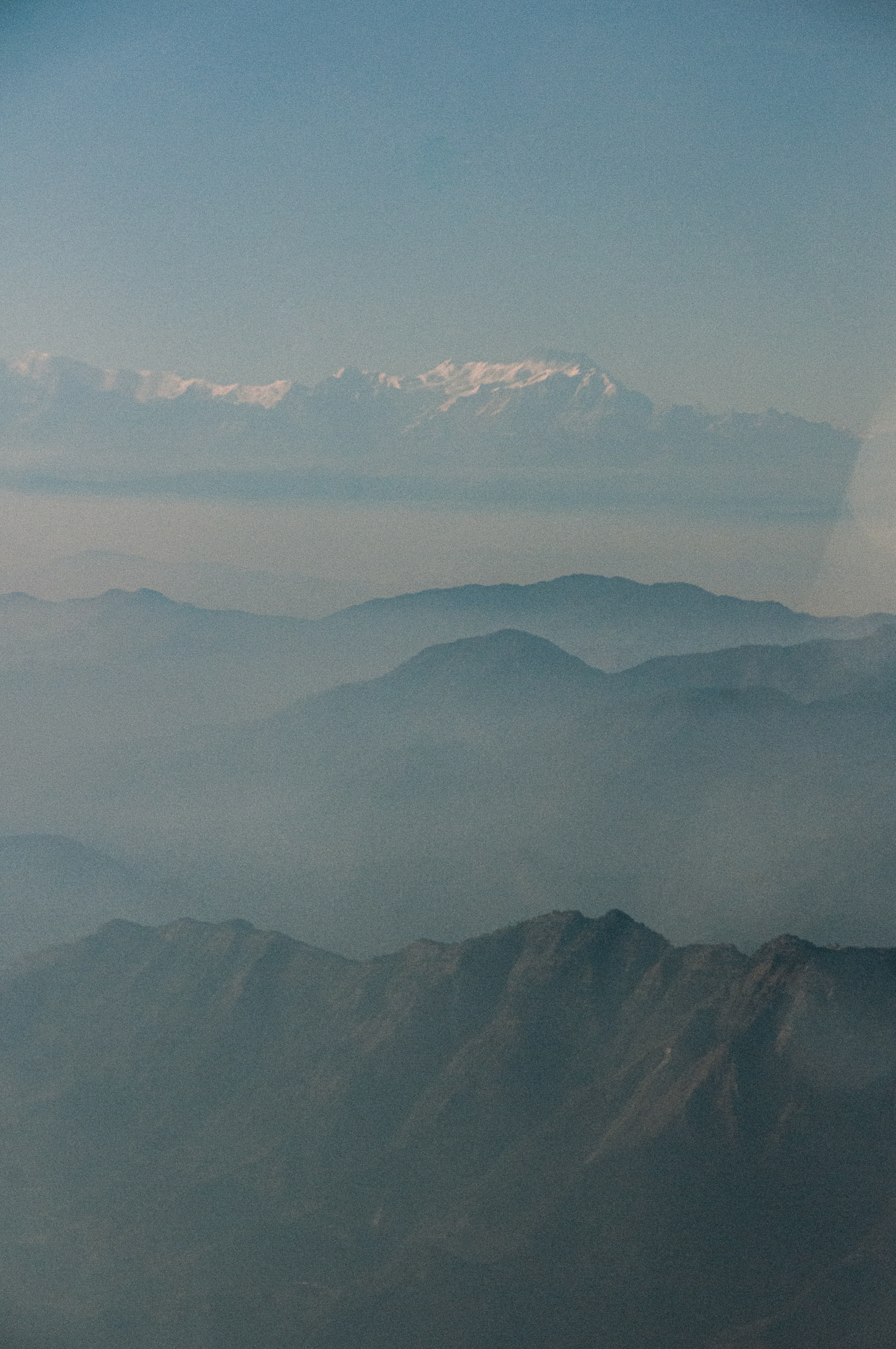 Multiple layers of dark mountain ridges fading into fog beneath a clear blue sky with distant snow-capped peaks.