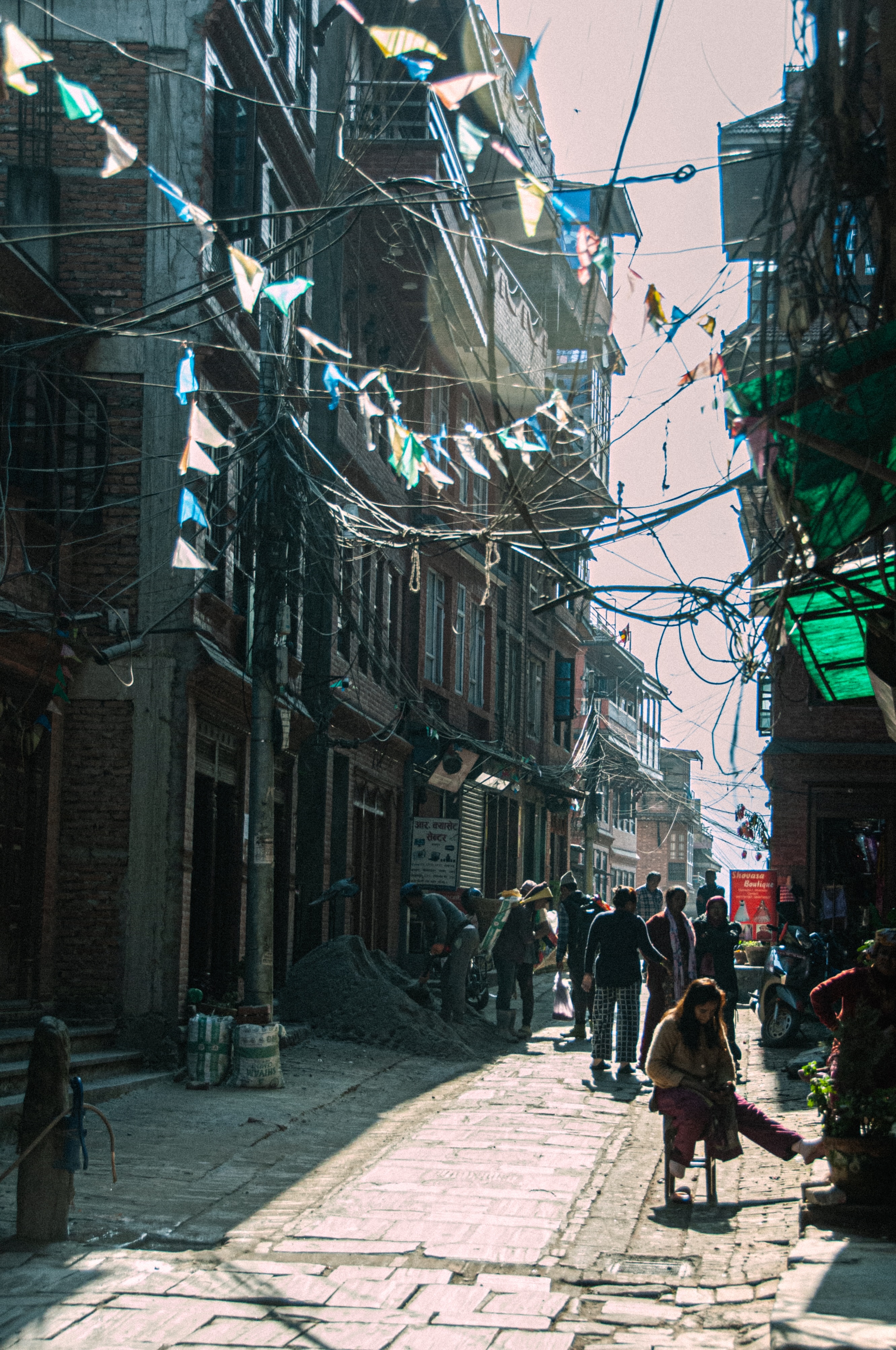 Narrow street with brick buildings, overhead electrical wires and festive flags, people walking and sitting on the sunlit stone pavement.