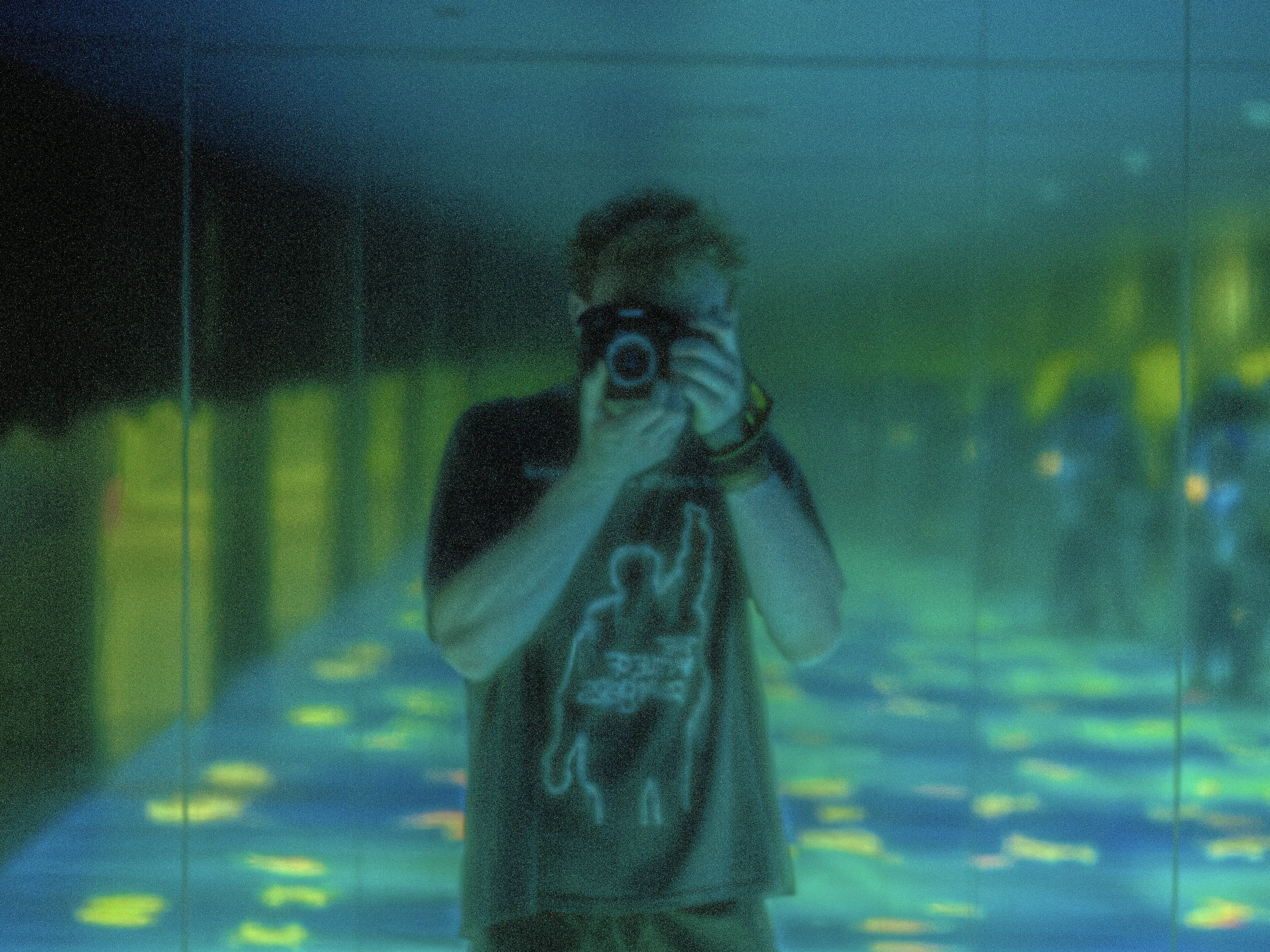 Person taking a photo in a reflective, dimly lit room with blue and green glowing floor patterns.