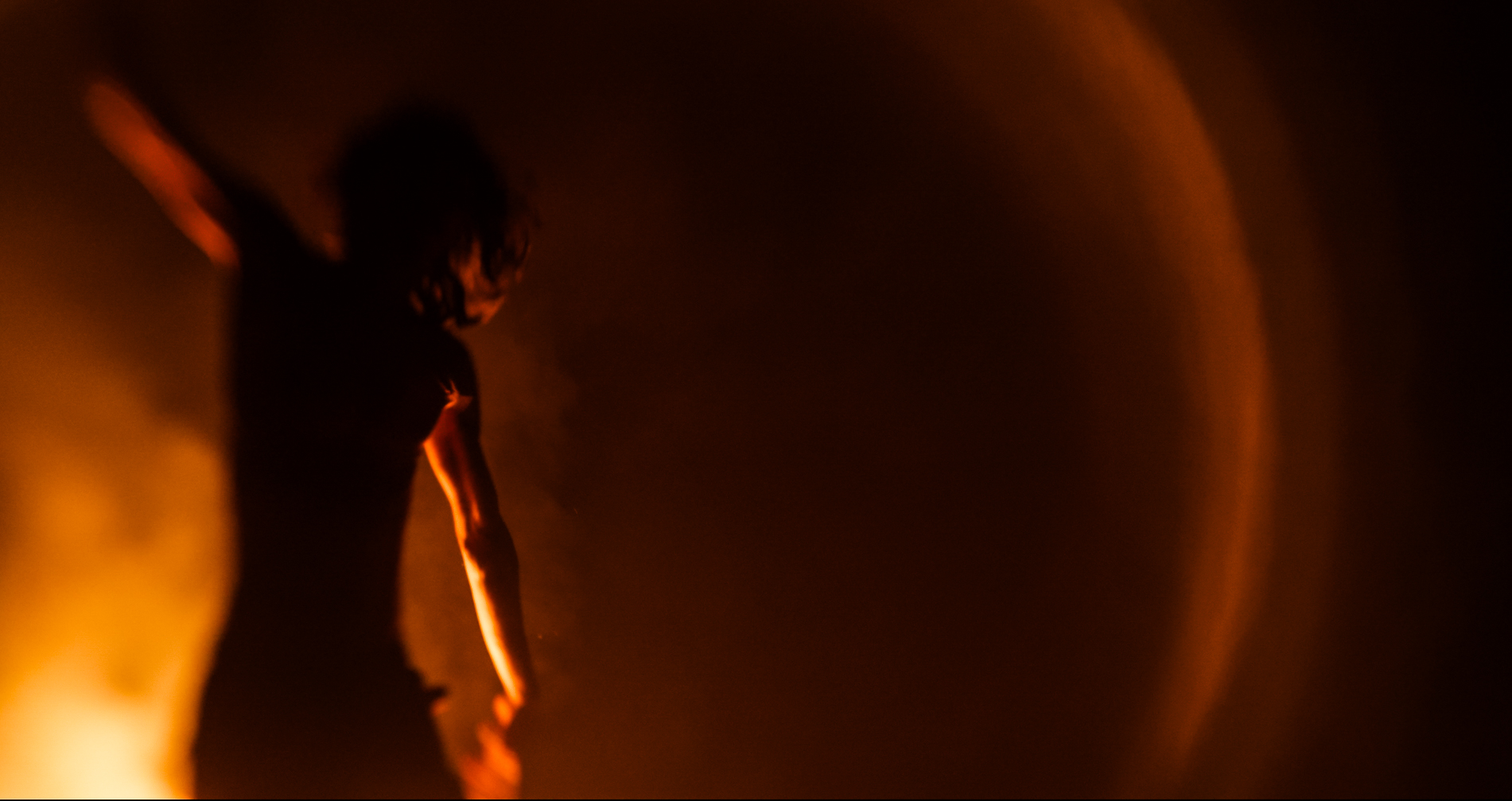 Silhouette of Tikaram dancing with an arm raised, illuminated by warm orange light and smoke.