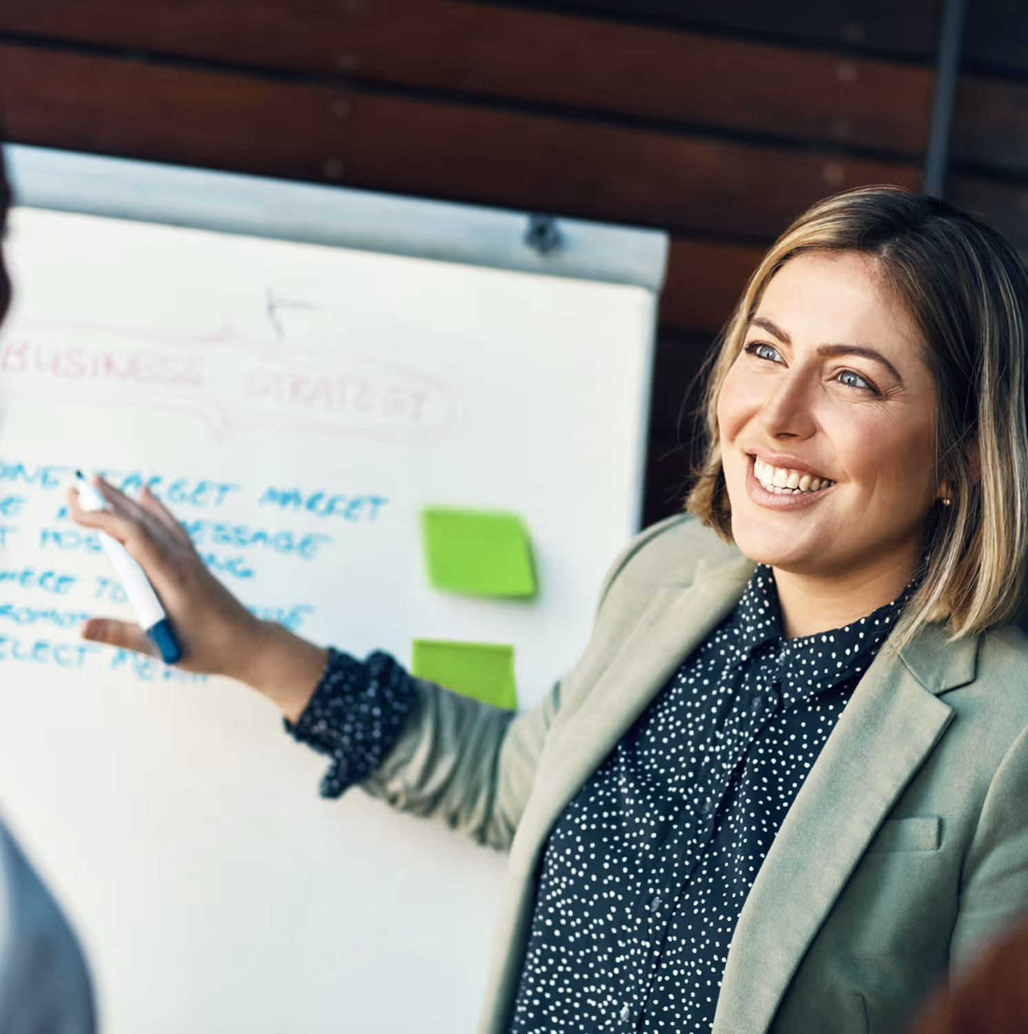 Frau mit hellbraunen Haaren erklärt lachend eine Geschäftsstrategie auf einem Whiteboard mit Haftnotizen.
