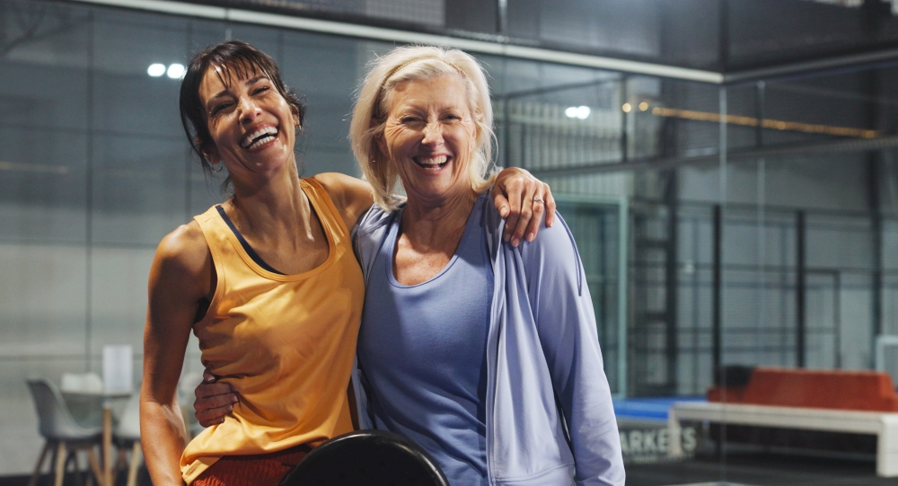 Two women smiling and embracing each other in a gym setting, one wearing an orange tank top and the other in a blue shirt and jacket.