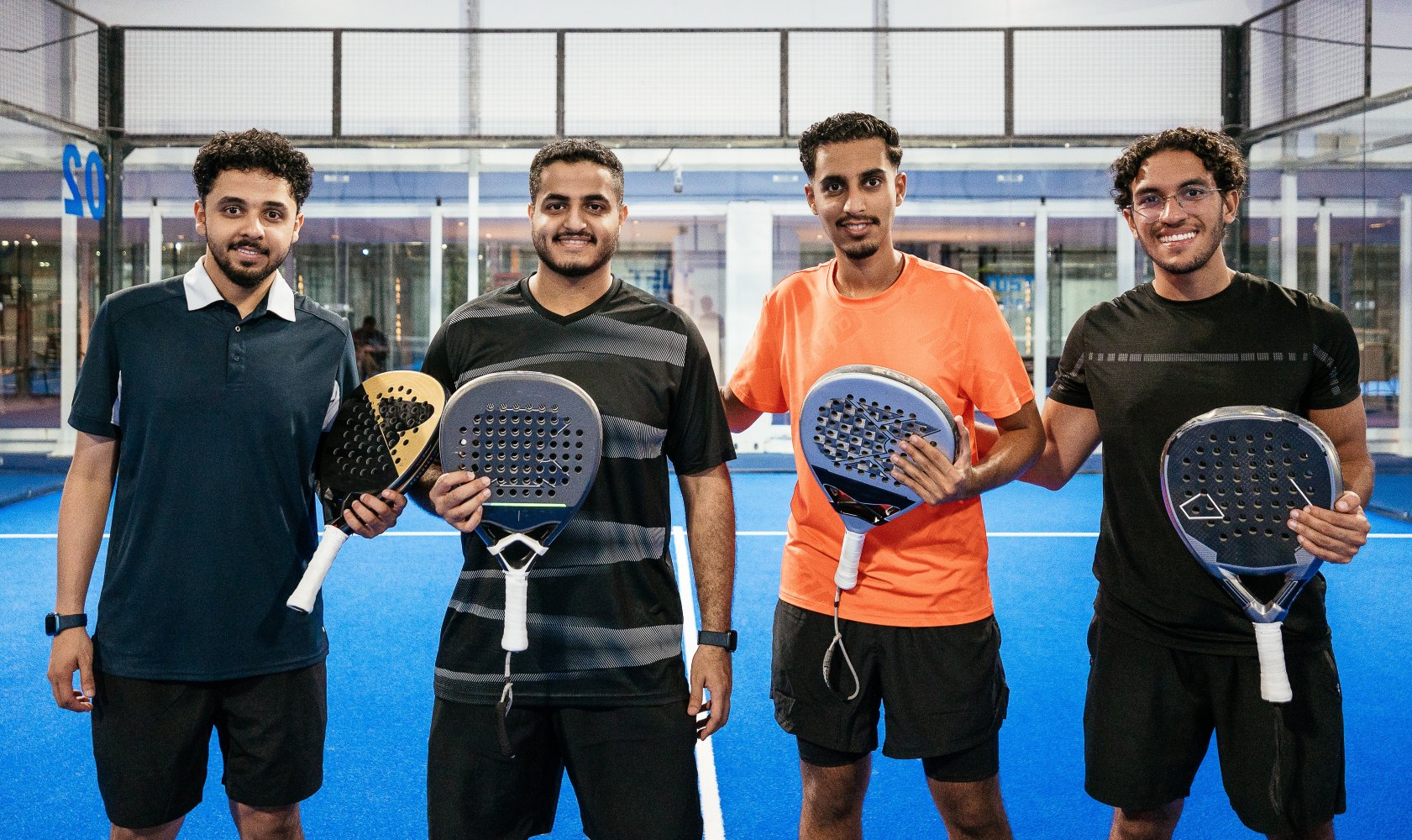 Four young men standing on a blue paddle tennis court holding paddles and smiling at the camera.