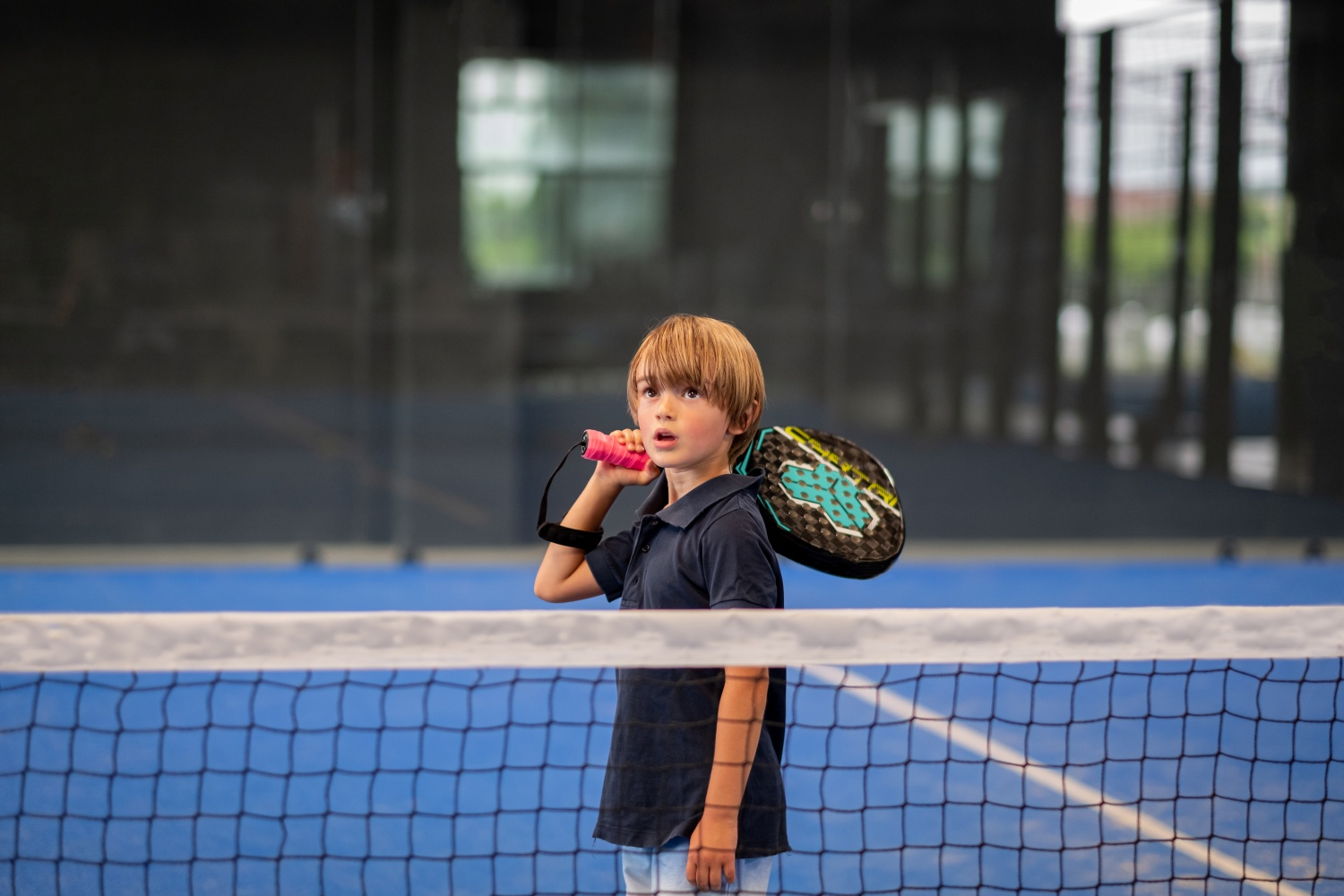 Young boy holding a paddle on his shoulder standing on a blue indoor paddle tennis court behind the net.