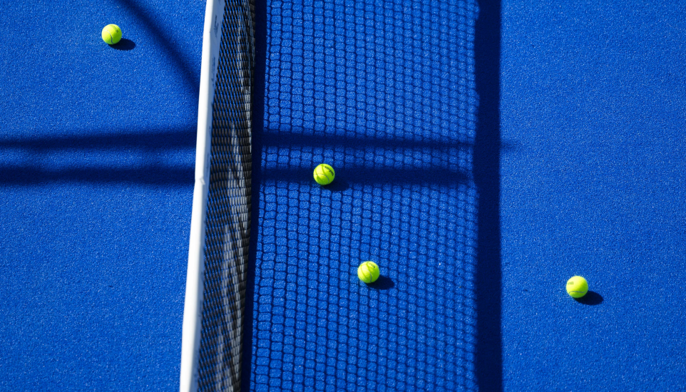 Four tennis balls on a bright blue tennis court with the net casting a shadow across the surface.