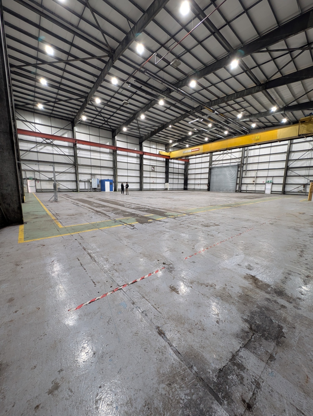 Spacious empty industrial warehouse with high metal ceiling, overhead crane, marked concrete floor, and two people standing in the distance.