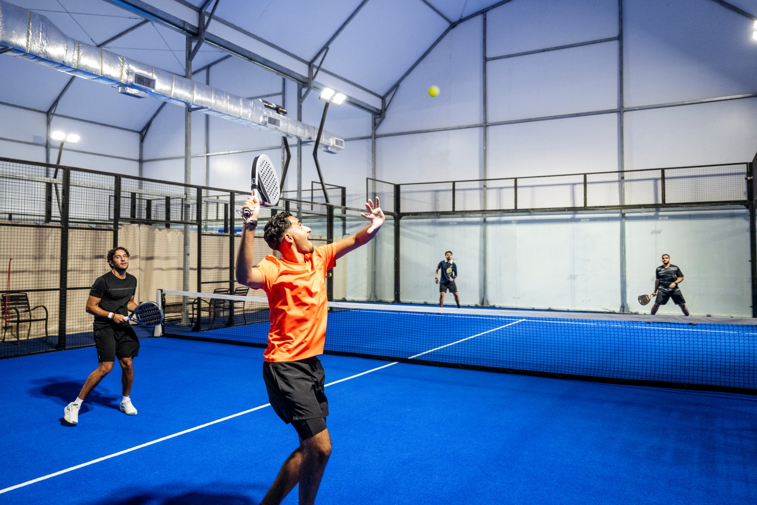 Four men playing padel tennis on a blue indoor court, with one in an orange shirt preparing to serve the ball.