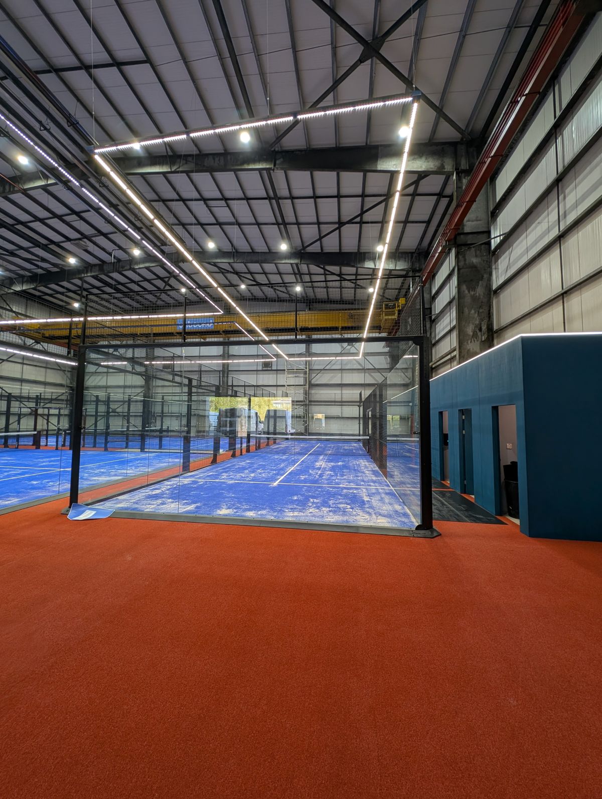 Illuminated padel tennis court with blue floor, black mesh fencing, and rectangular overhead LED lights in a dark environment.