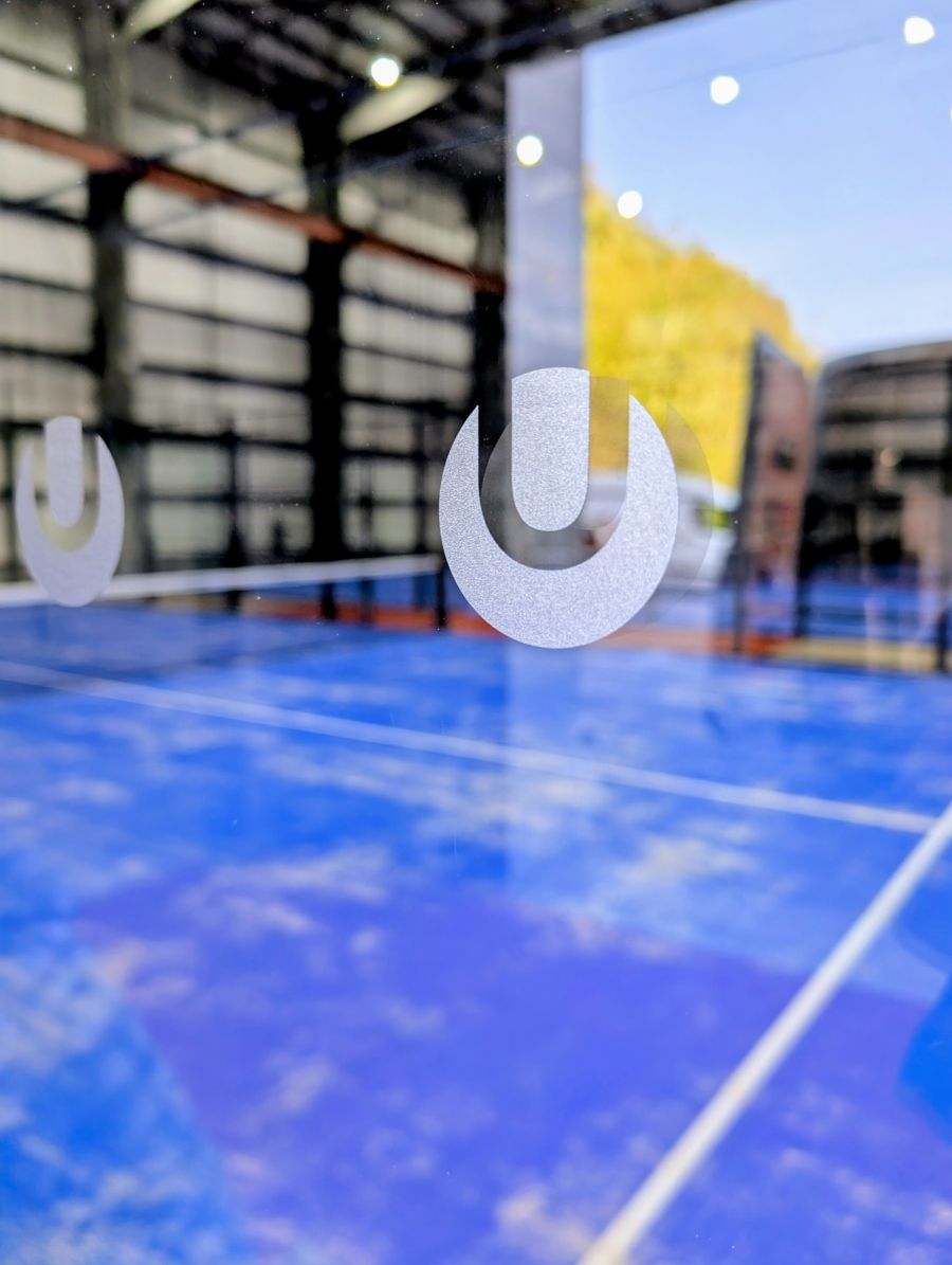 Indoor padel tennis court with blue flooring and glass walls under artificial lighting.