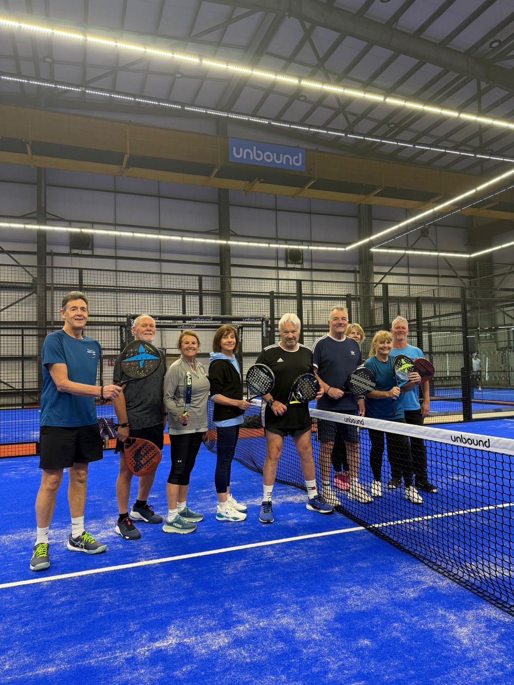 Eight adults standing on an indoor blue padel court holding padel rackets near the net under 'unbound' sign.