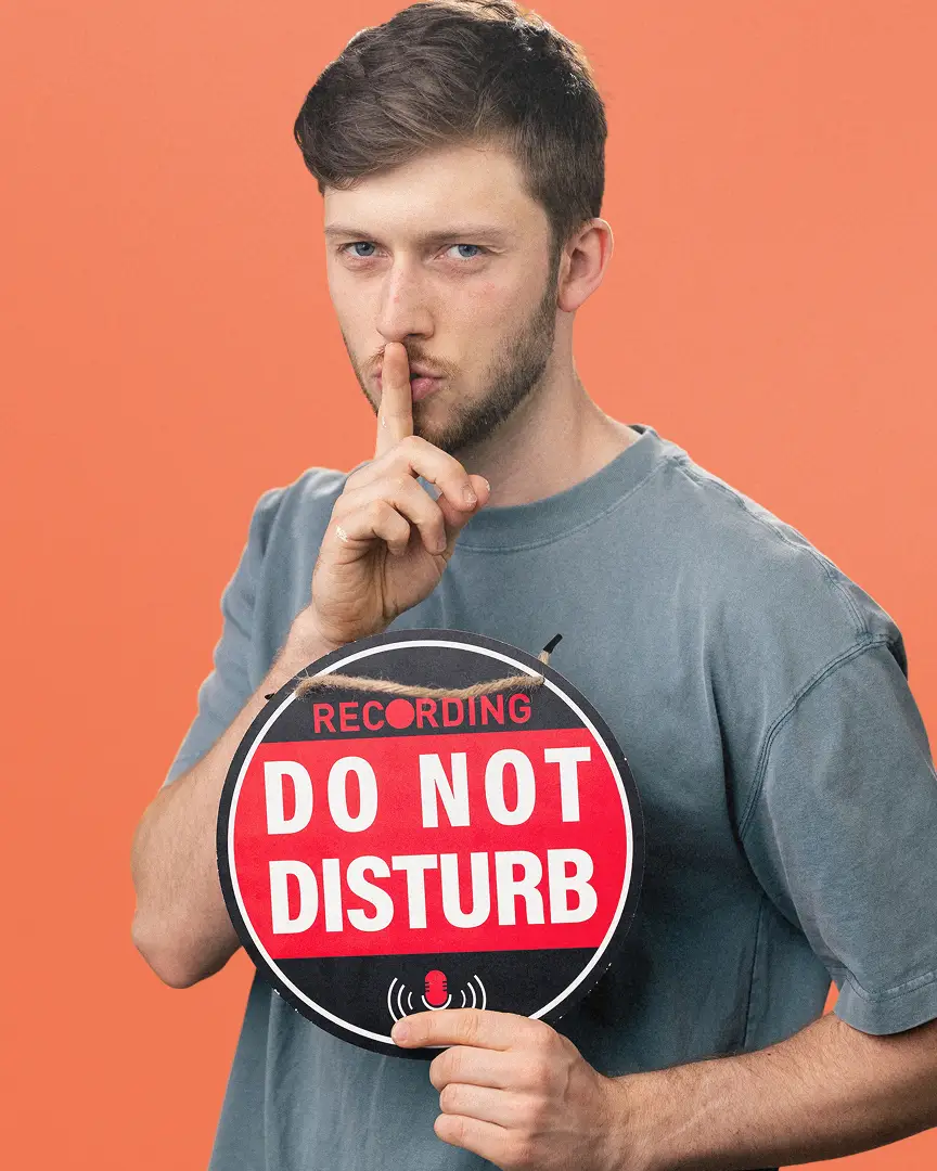 Ben holding a round sign that says 'Recording Do Not Disturb' with his finger to his lips signaling silence.