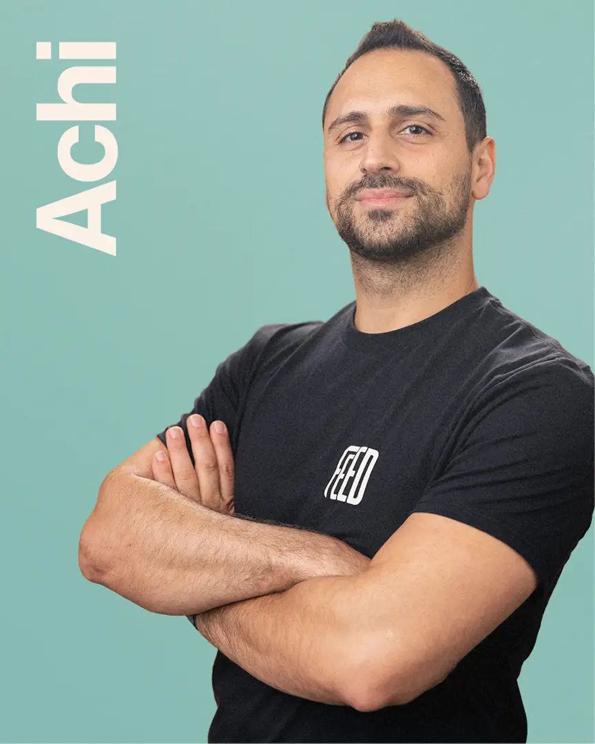 Achi wearing a black FEED t-shirt, standing with arms crossed with the word 'Achi' in vertical text.