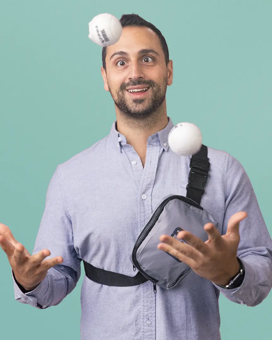 Achi wearing a light blue shirt and chest bag juggling two white balls.