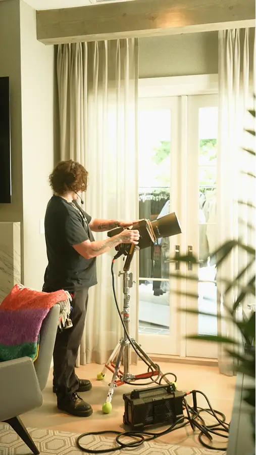 Person adjusting studio light on a stand in a bright room with large windows and curtains.